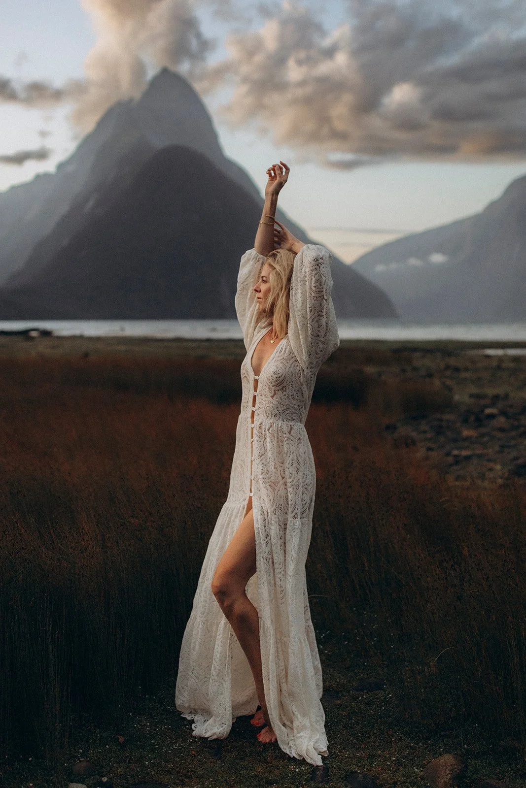 A woman in a white lace dress standing outdoors with arms raised, against a backdrop of mountains and cloudy sky.