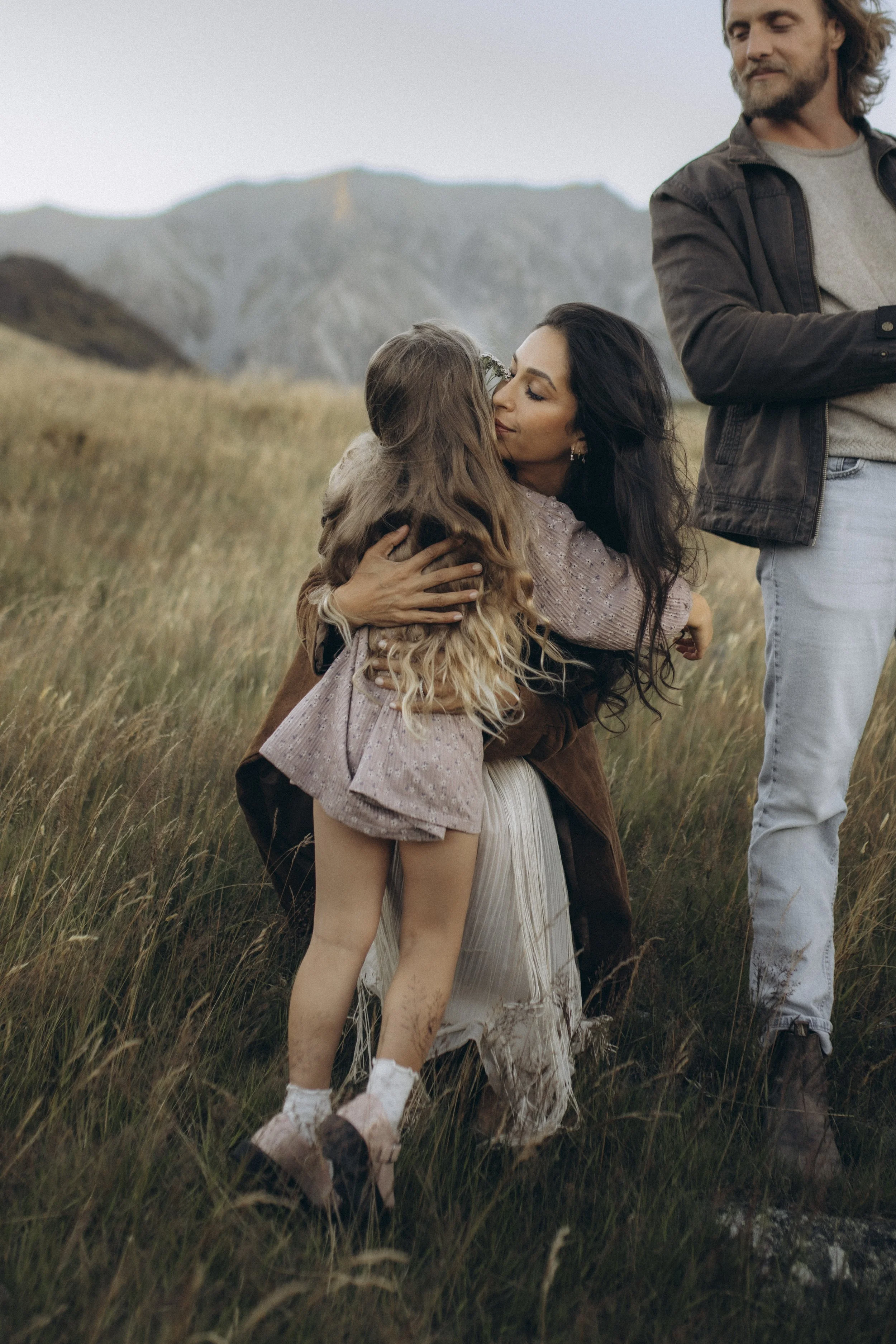 A woman with dark hair wearing a light pink dress hugging a young girl with long hair and a pink dress in a field of tall grass with mountains in the background, accompanied by a man with long hair and a beard standing nearby.
