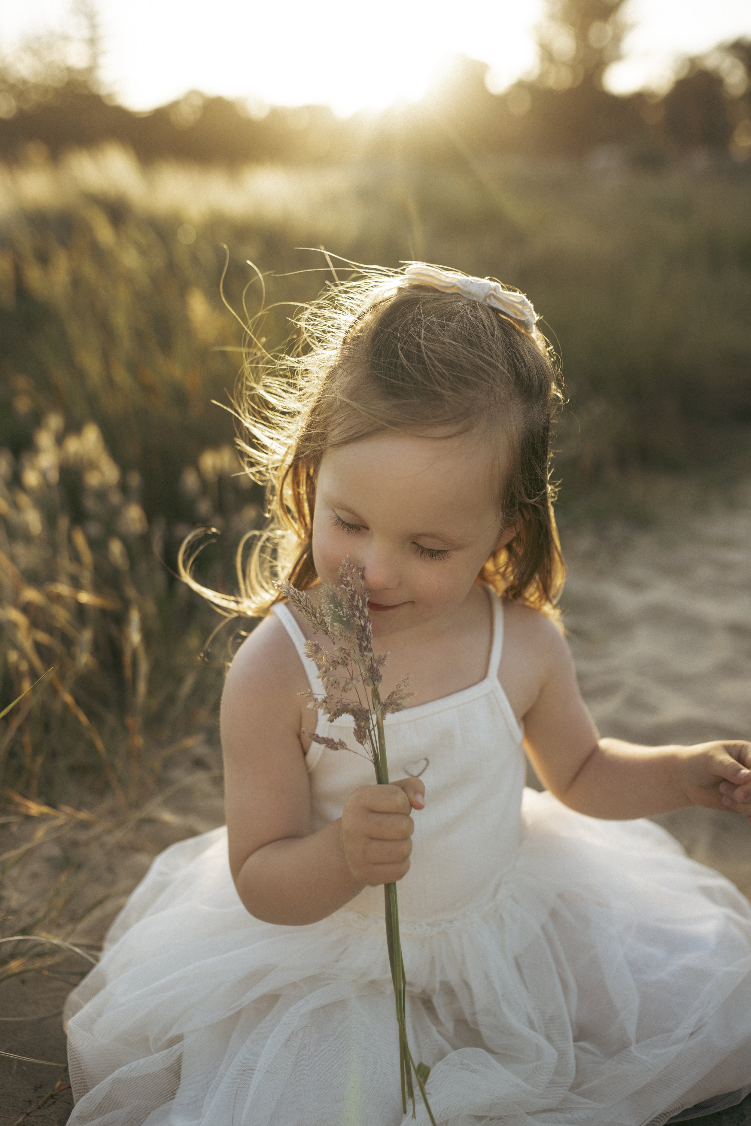 A young girl with red hair and a white headband, sitting outdoors in a white dress, holding a small bouquet of dried flowers in her hand, with her eyes closed and a gentle smile, during sunset in a field of tall grasses.
