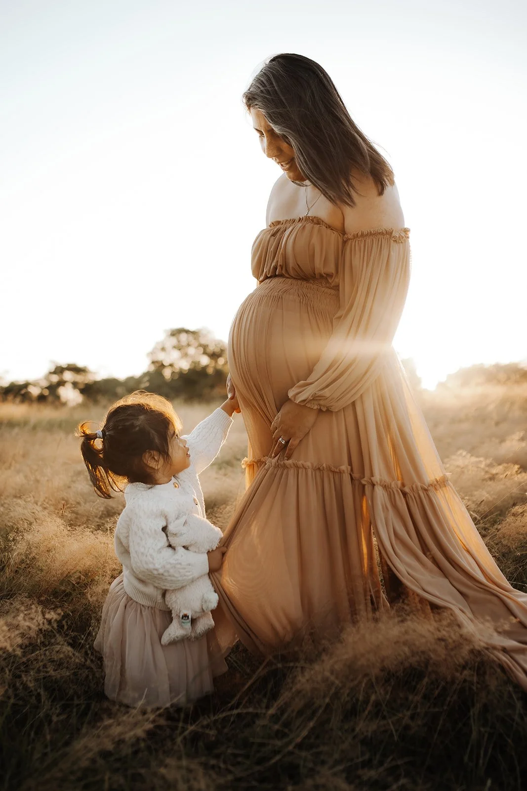 A pregnant woman in a flowing beige dress standing in a field at sunset, with a young girl touching her belly, holding a teddy bear, and looking up at her.