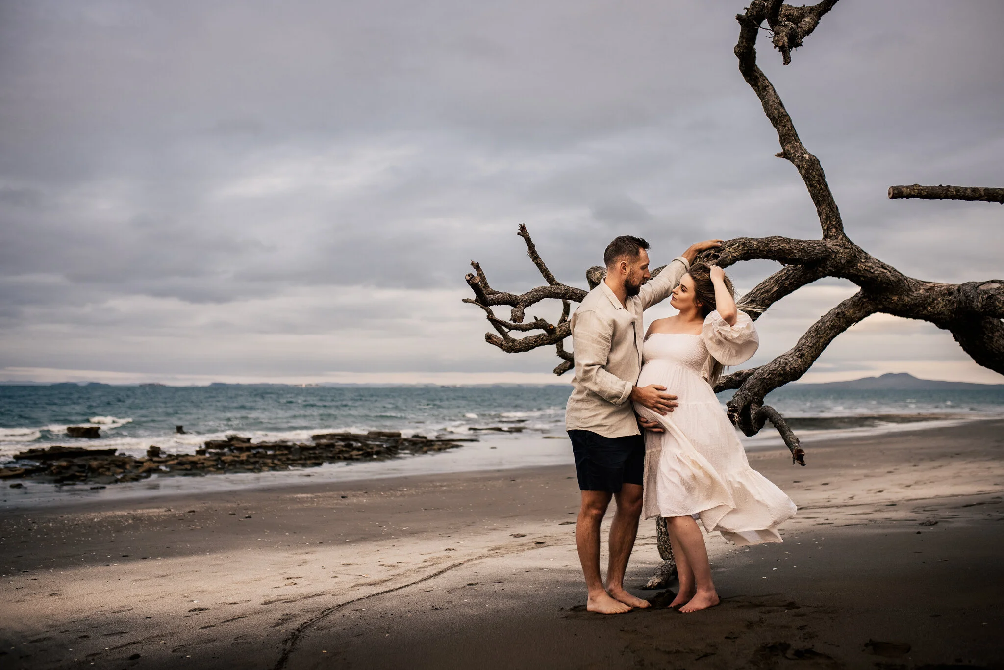A couple standing on a beach next to a large, twisted tree with the ocean and a cloudy sky in the background. The man is touching the woman's head, and she is pregnant, wearing a white dress.