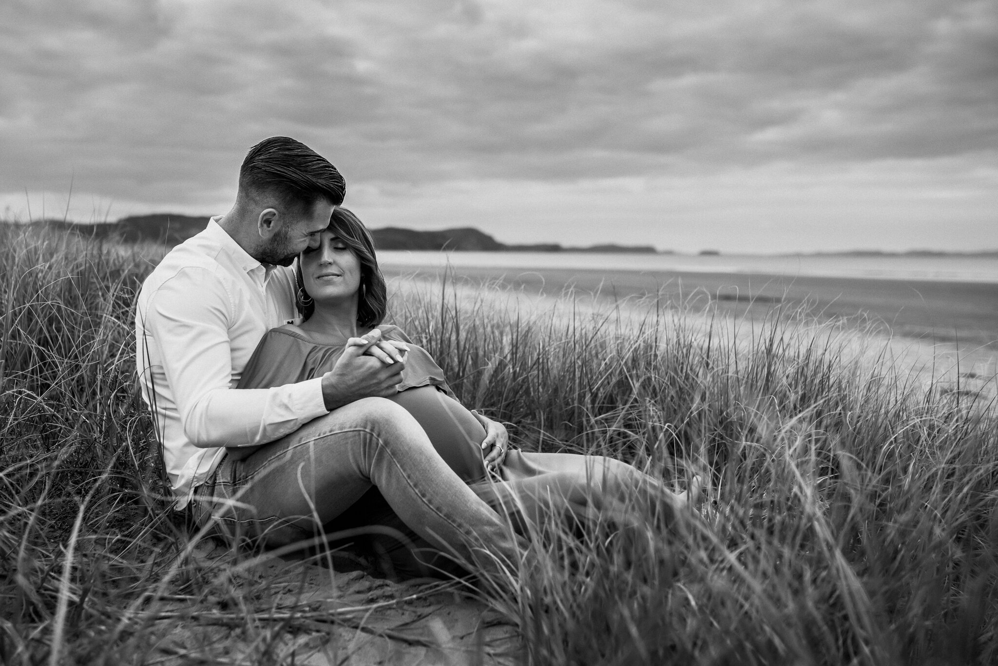 A black and white photo of a couple sitting on a beach near tall grass, with ocean waves and distant mountains under a cloudy sky in the background. They are embracing and holding hands, with the man resting his forehead against the woman's forehead,