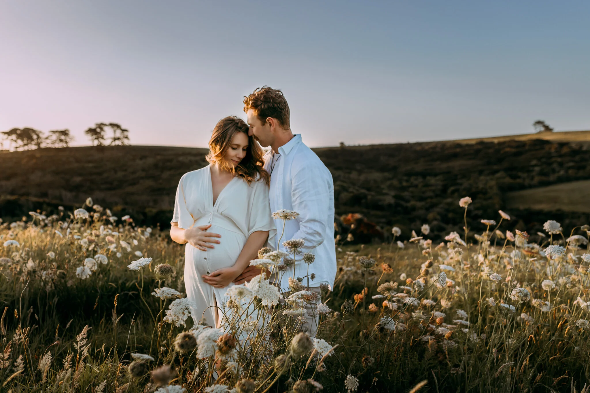 A pregnant woman and a man embracing in a field of flowers during sunset.