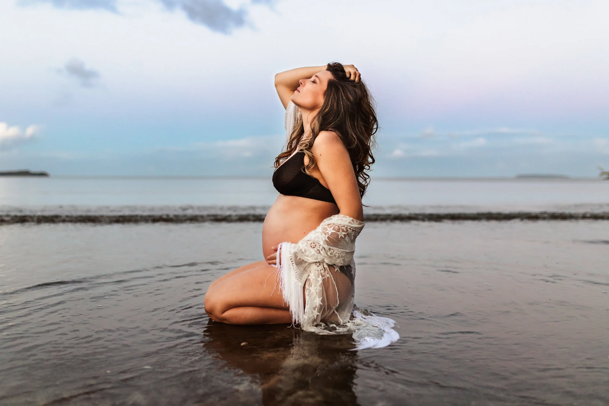 A pregnant woman kneeling in shallow water on a beach with her hand on her head, wearing a black swimsuit and white lace cover-up, with a cloudy sky in the background.