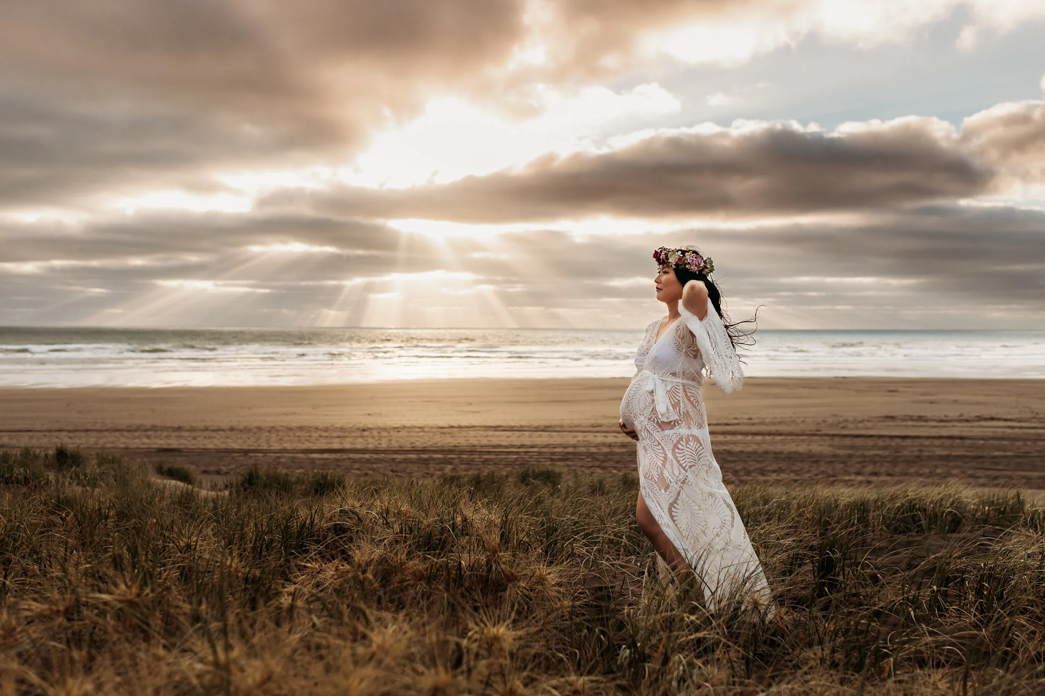 A pregnant woman wearing a white lace dress and floral crown standing on a beach with grassy dunes, ocean, and cloudy sky with sun rays in the background.