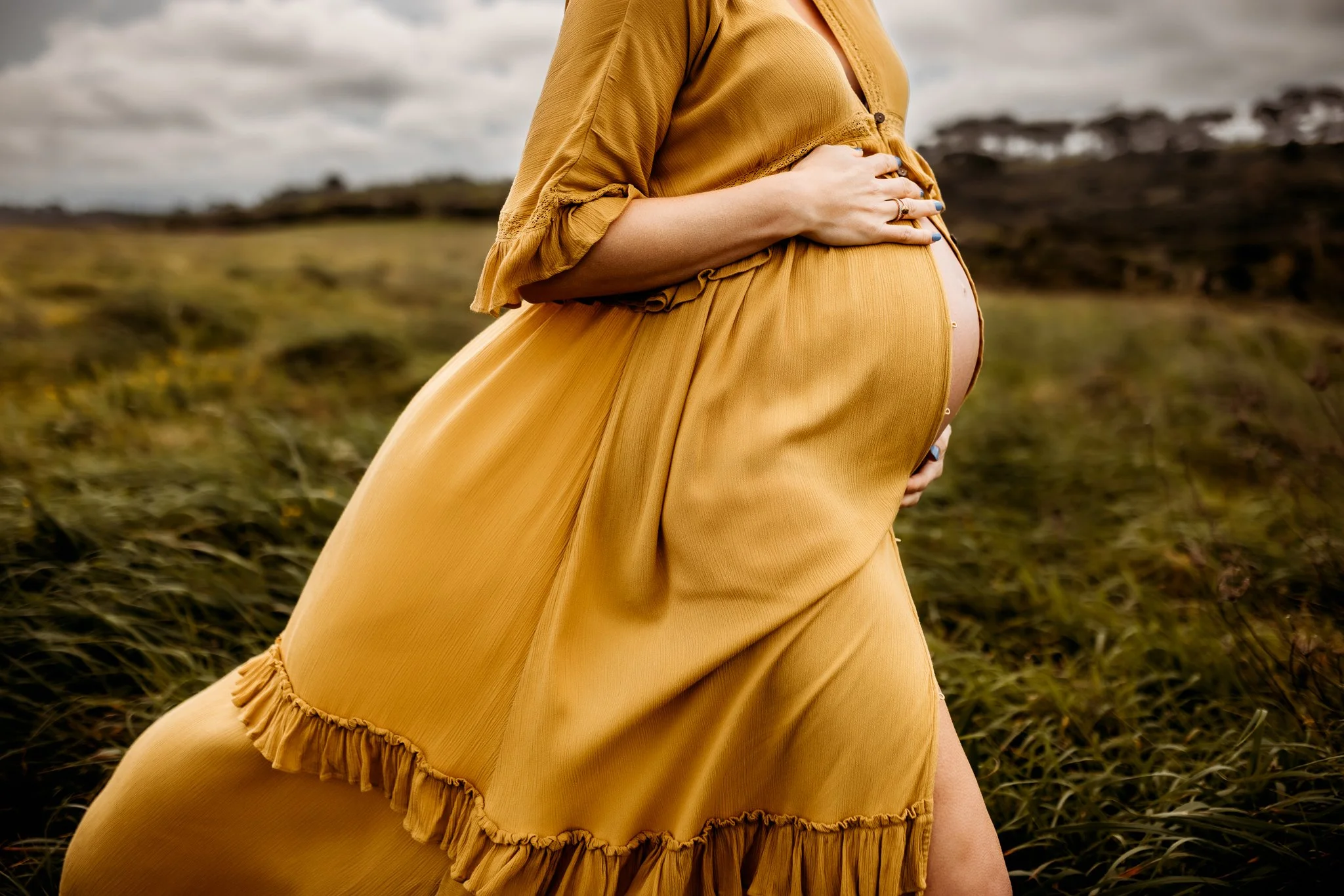 A pregnant woman wearing a mustard-colored dress with ruffled details, standing outdoors in a grassy field with cloudy skies.