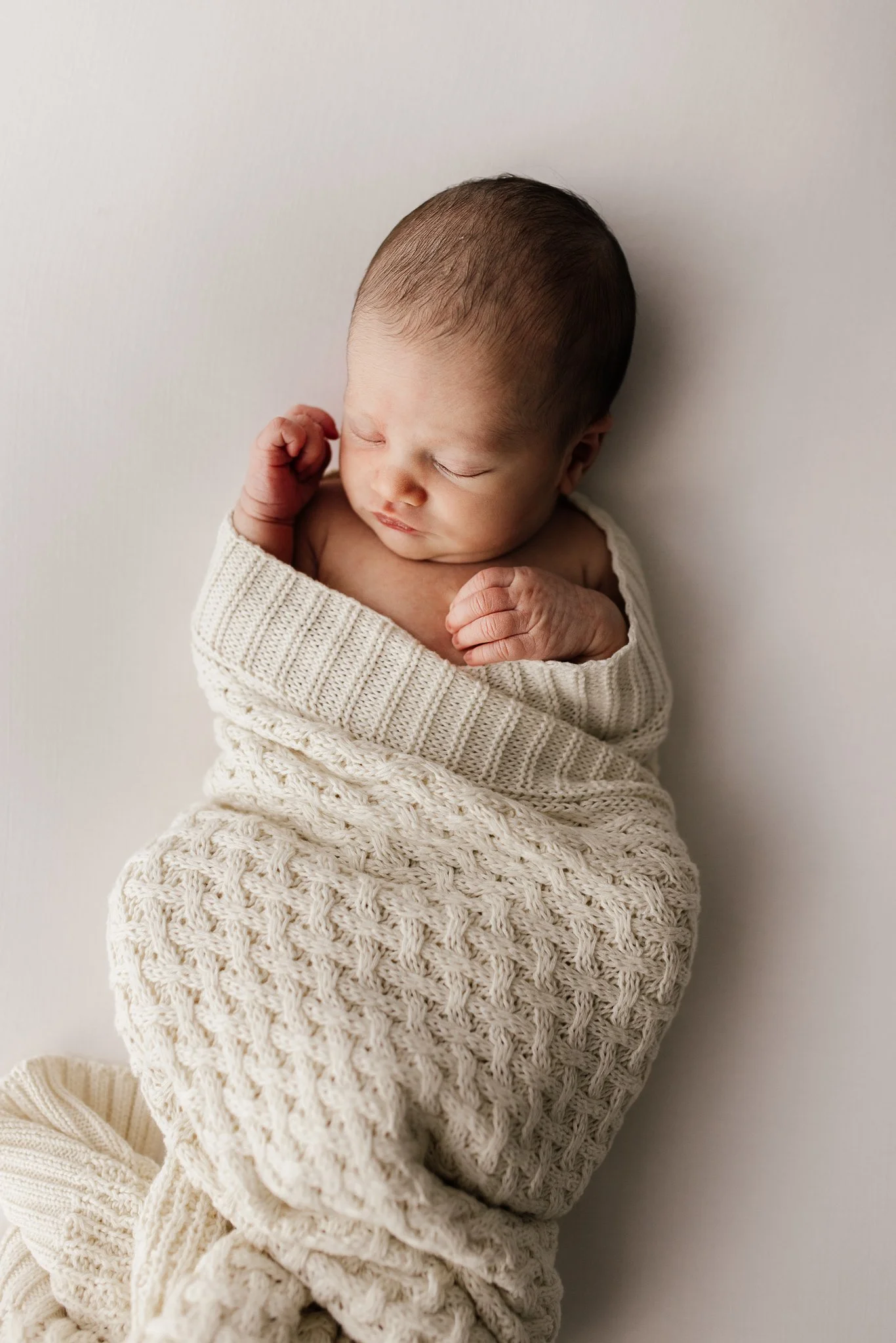 A newborn baby sleeping peacefully, wrapped in a cream-colored knitted blanket, with a matching knitted hat, laying on a plain white surface.