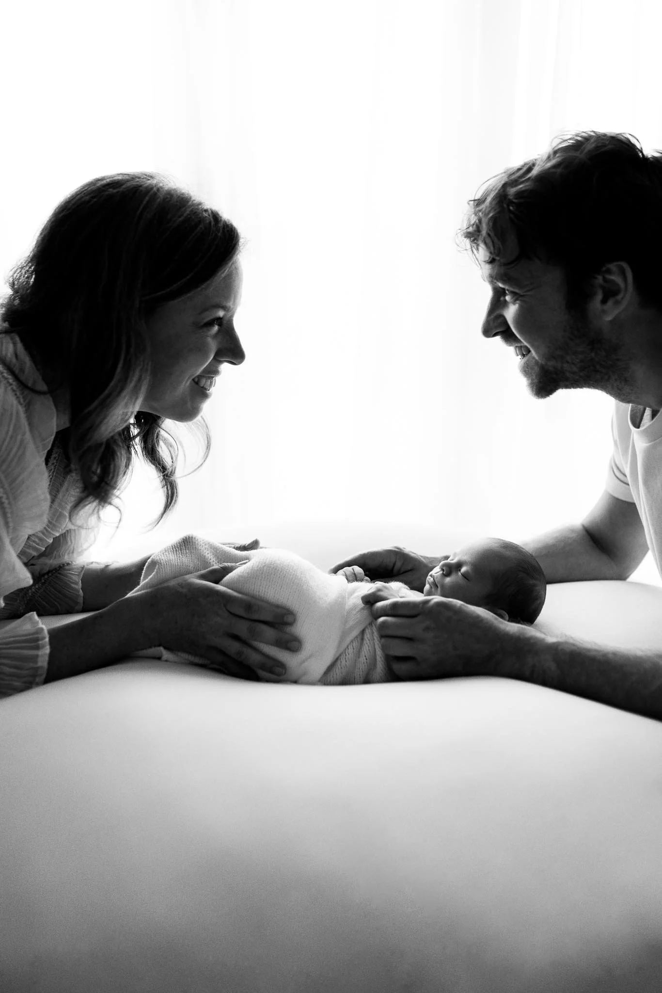 Black and white photo of a woman and a man smiling and leaning towards each other, holding a sleeping baby.