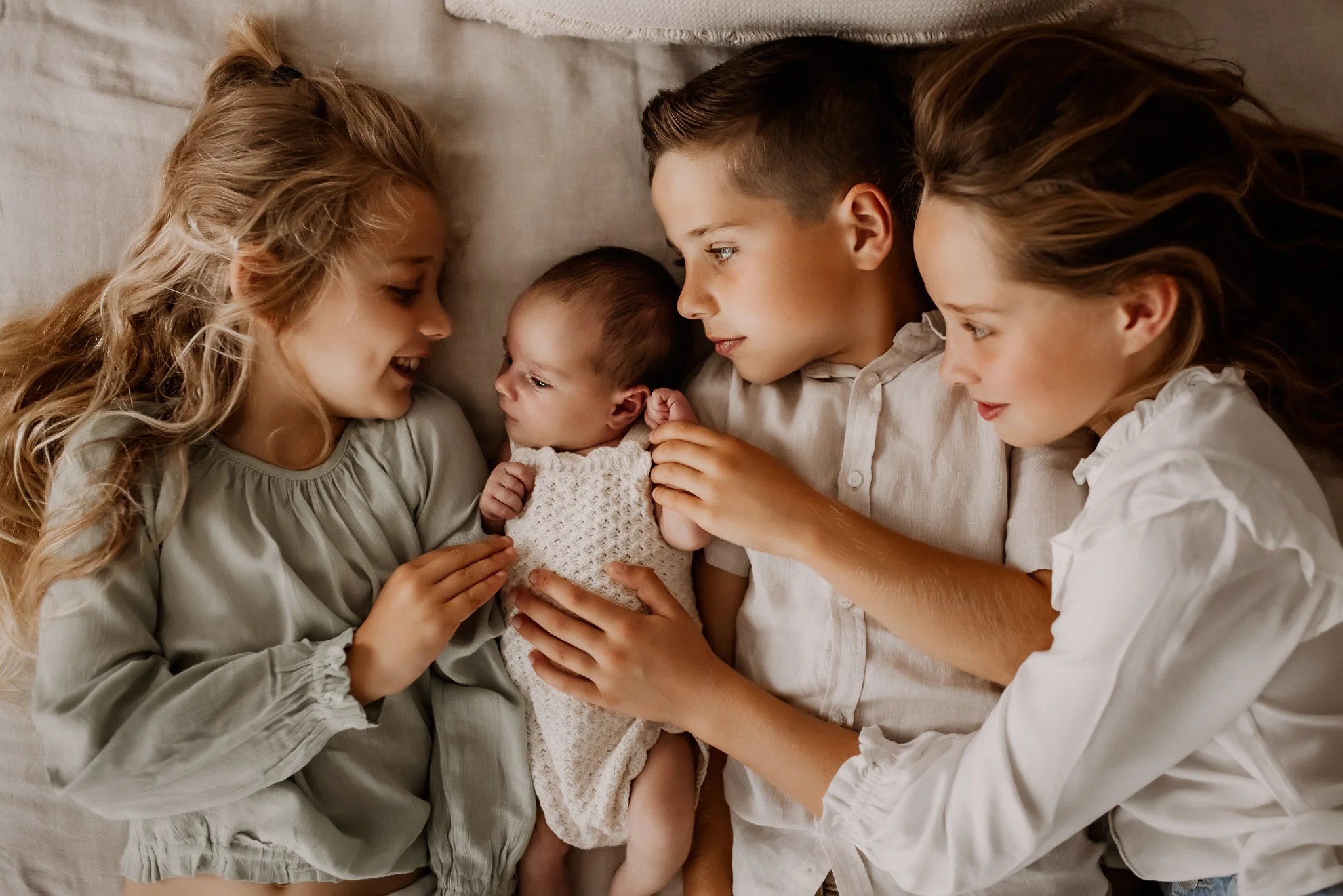 Four siblings lie close together on a bed, gently holding a newborn baby while looking at the baby with affection.