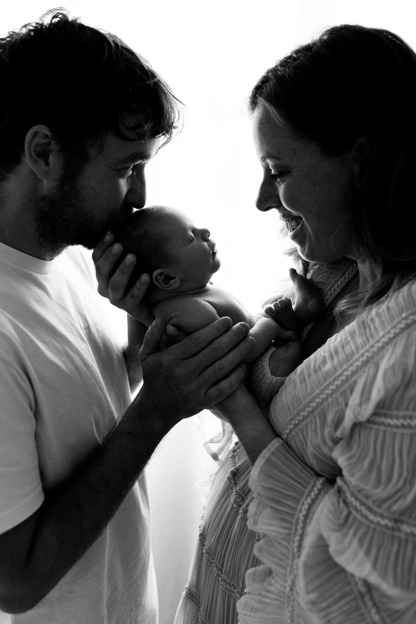 A black-and-white photo of a man and a woman holding a baby, with their faces close together, smiling and touching foreheads in a tender moment.