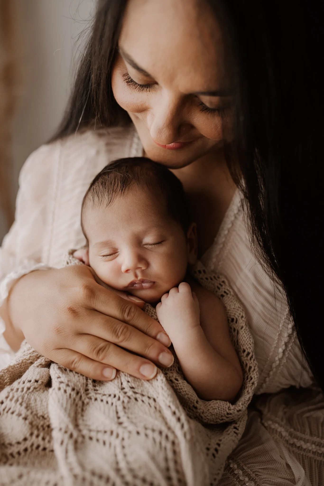 A woman with long dark hair holding a sleeping baby wrapped in a beige knitted blanket.