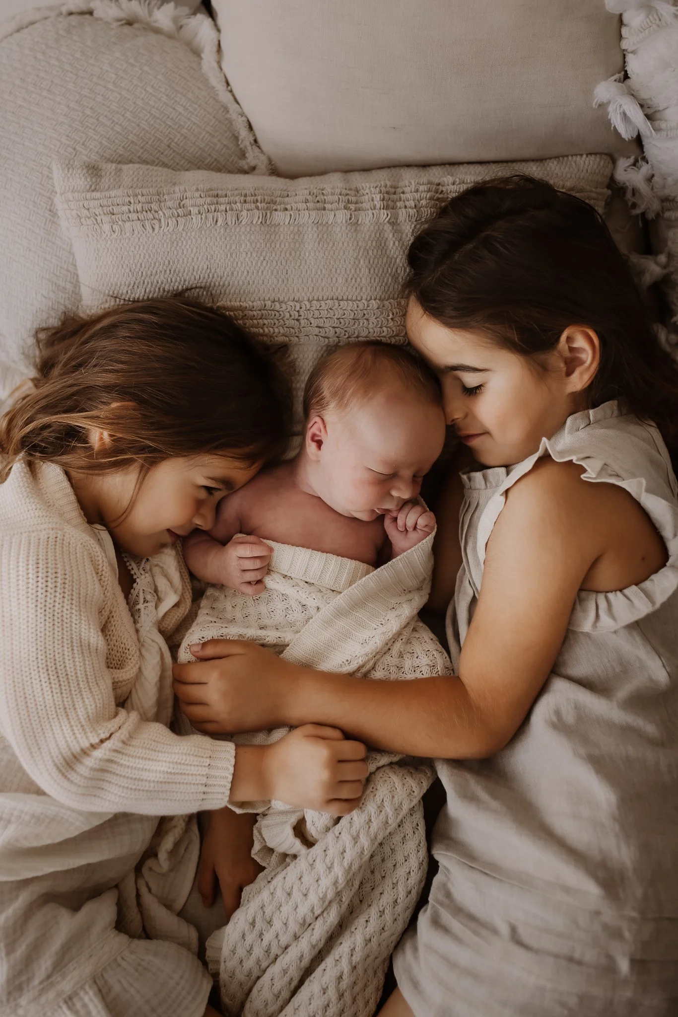A group of three children, an infant and two girls, lie close together on a bed with neutral-colored, textured blankets and pillows, cuddling peacefully.