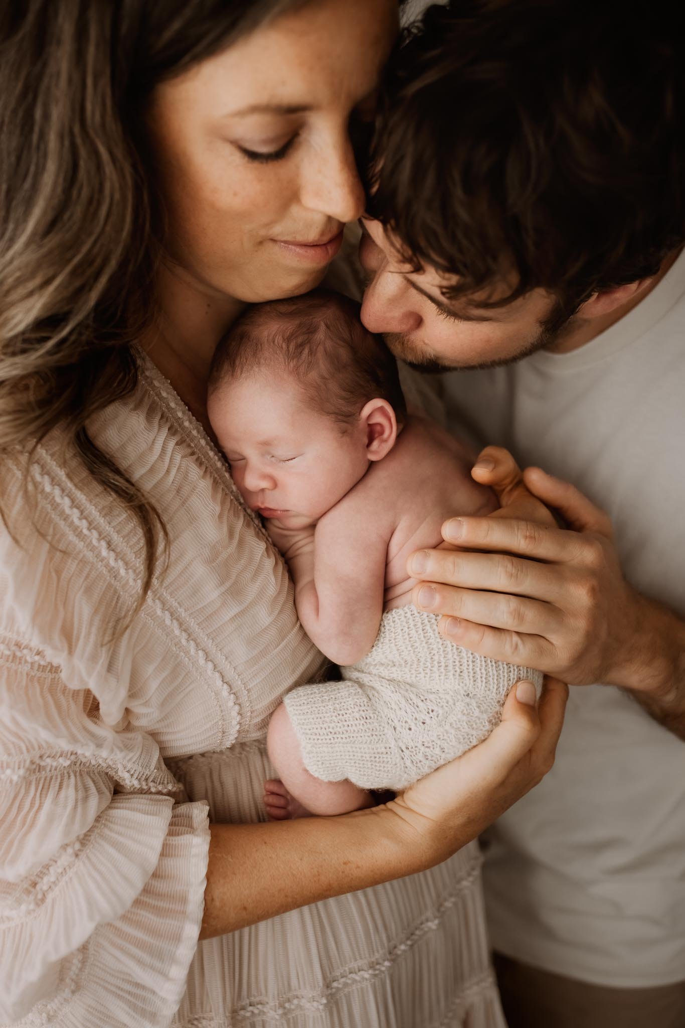 A woman and a man are holding a newborn baby close to her chest. The woman has long brown hair and is wearing a beige, textured dress. The man has dark curly hair, a beard, and is wearing a white shirt. They are all gently touching their foreheads an