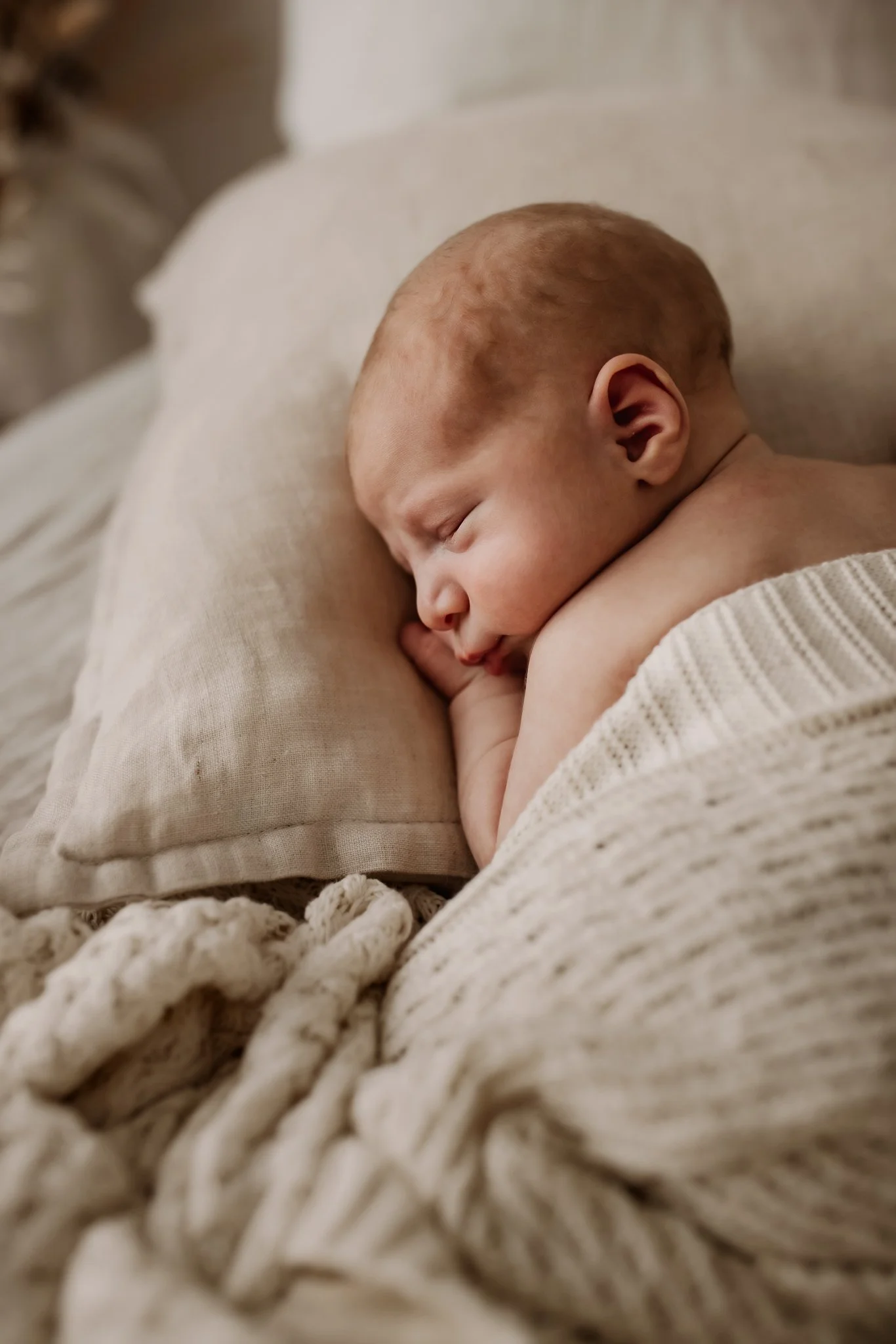 Close-up of a sleeping baby with reddish hair lying on a soft, cream-colored blanket and resting on a beige cushion.