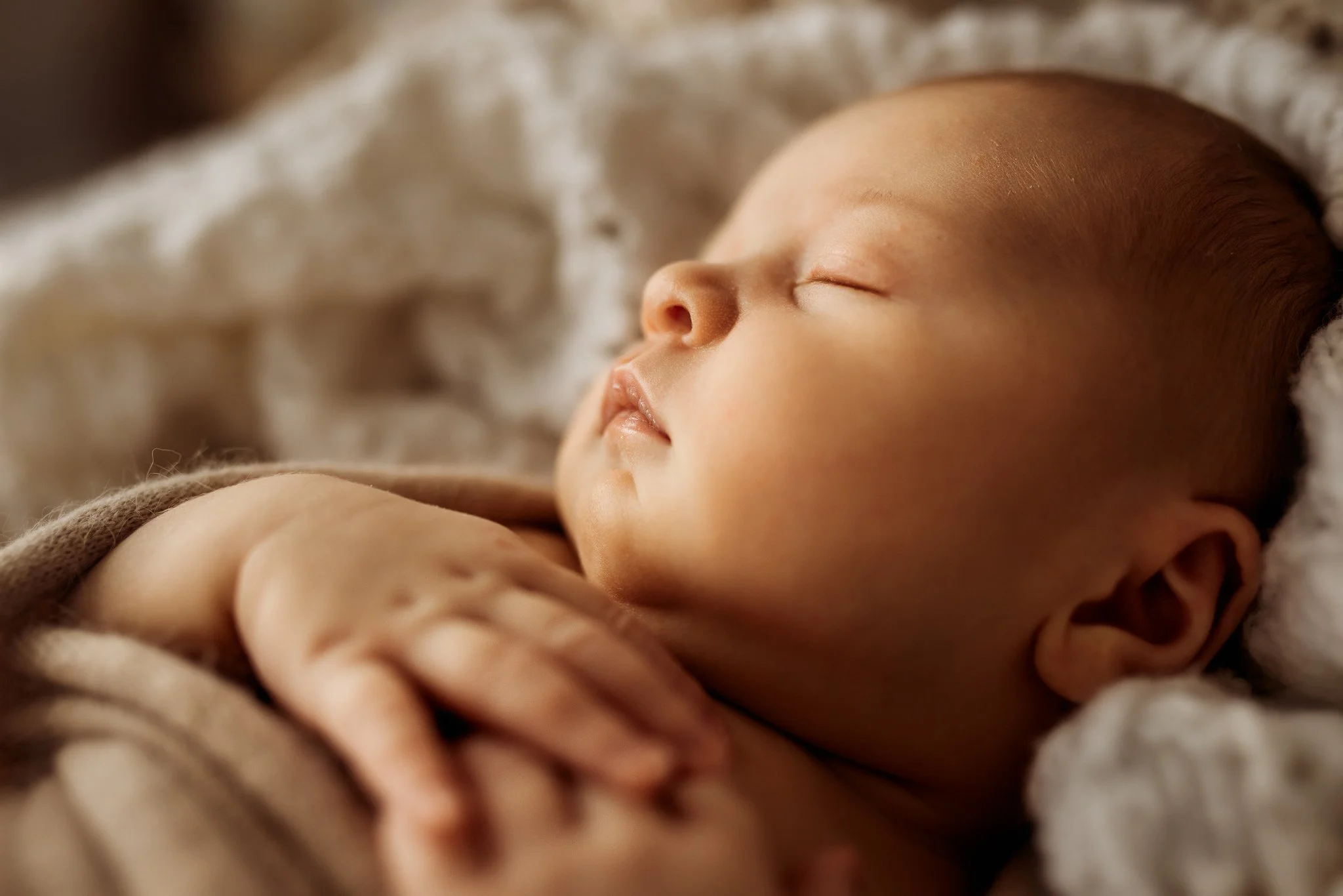 Close-up of a sleeping newborn baby's face and hand, lying on a soft, textured blanket.