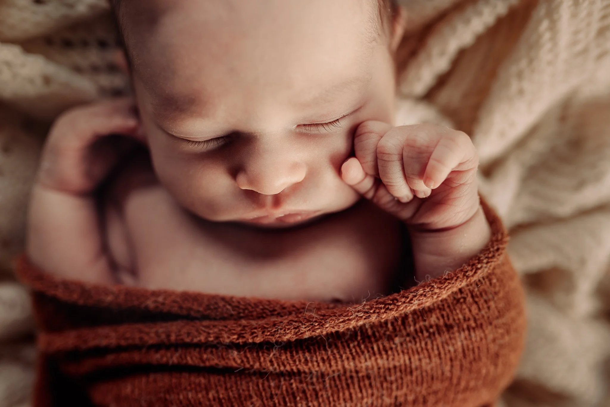 Close-up of a sleeping baby with closed eyes, hand near face, wrapped in a soft, warm, brown knitted blanket.