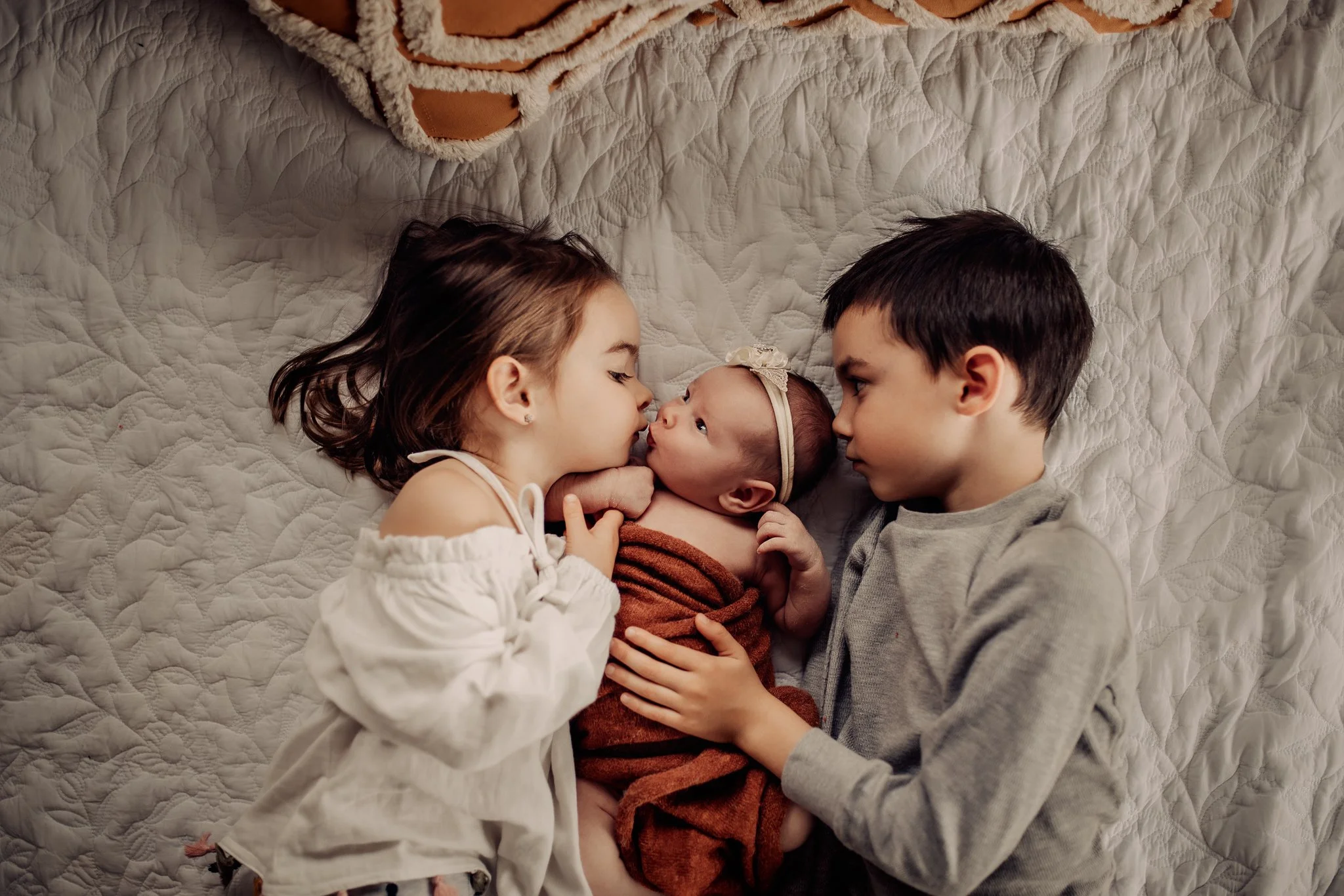 A young girl, a baby, and a boy are lying on a quilted bed, facing each other, with the girl and boy touching noses and the baby in the middle, looking at the girl.