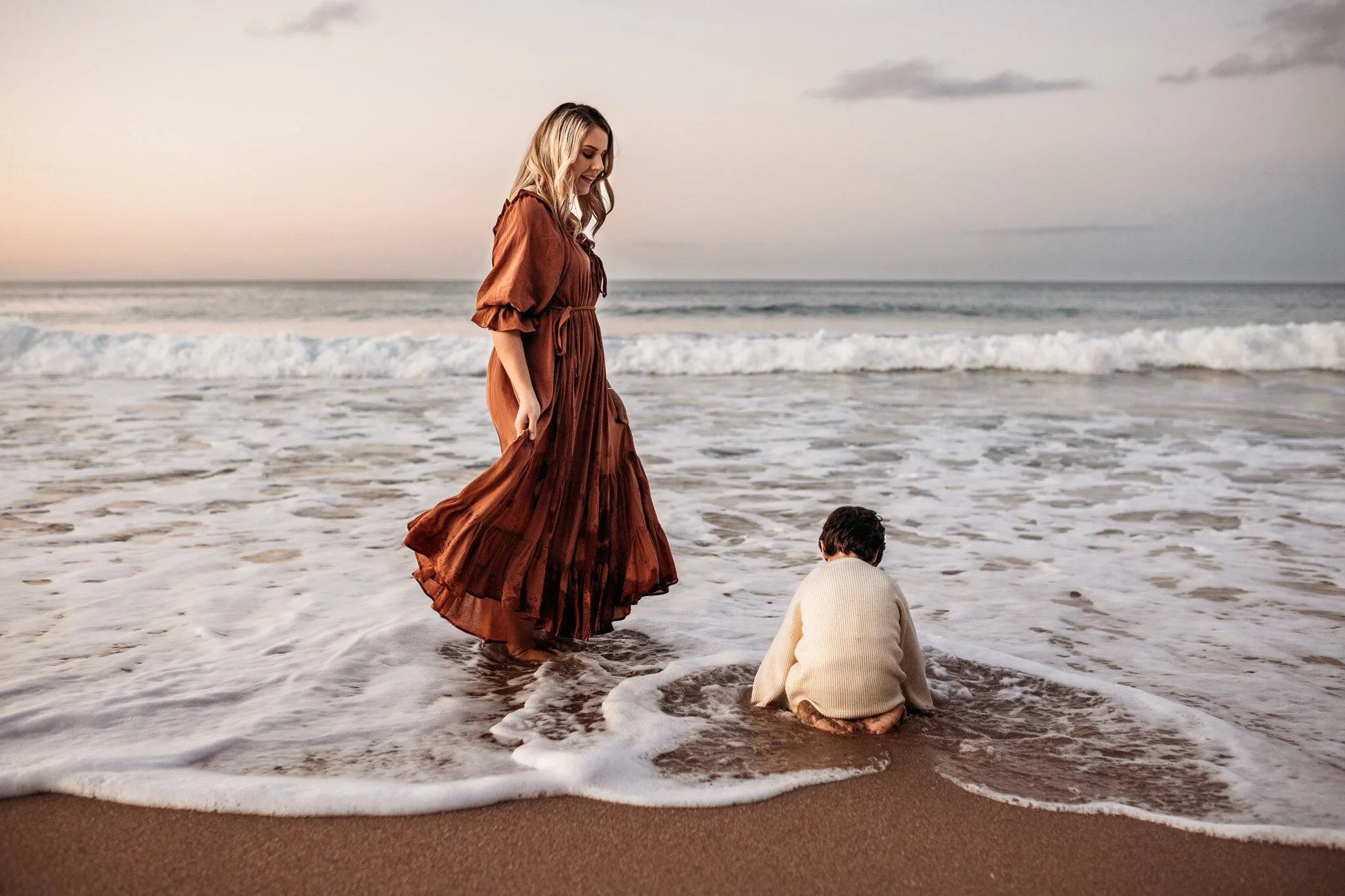 A woman in a long brown dress standing at the edge of the ocean, looking down at a child sitting in the sand by the water, with waves and cloudy sky in the background.