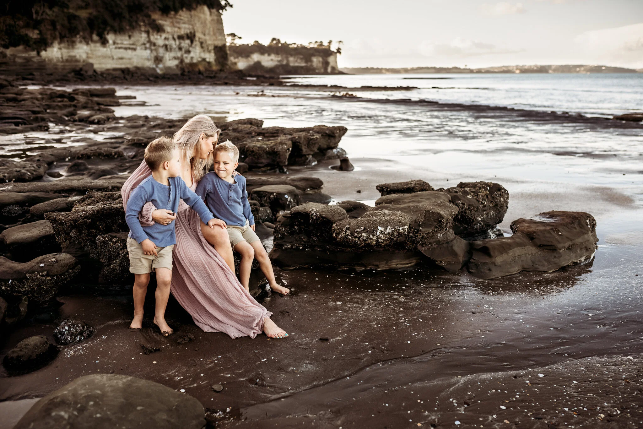 A woman with two young boys sitting on rocks at the beach during sunset, with cliffs and water in the background.