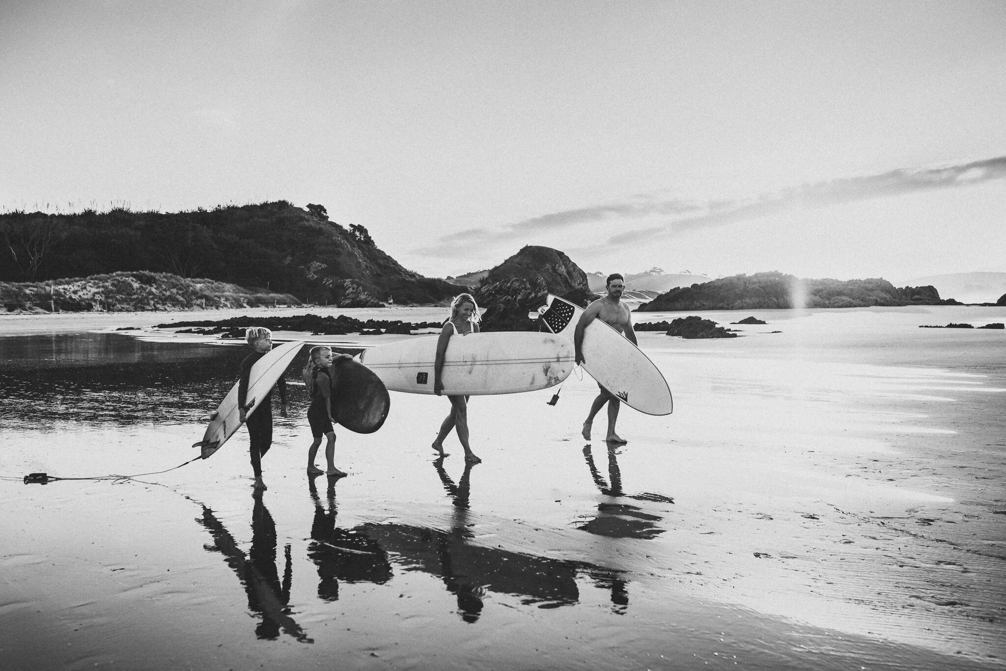 A group of five people, including children, carrying surfboards and a ball, walking along the beach near the water with rocky hills in the background.