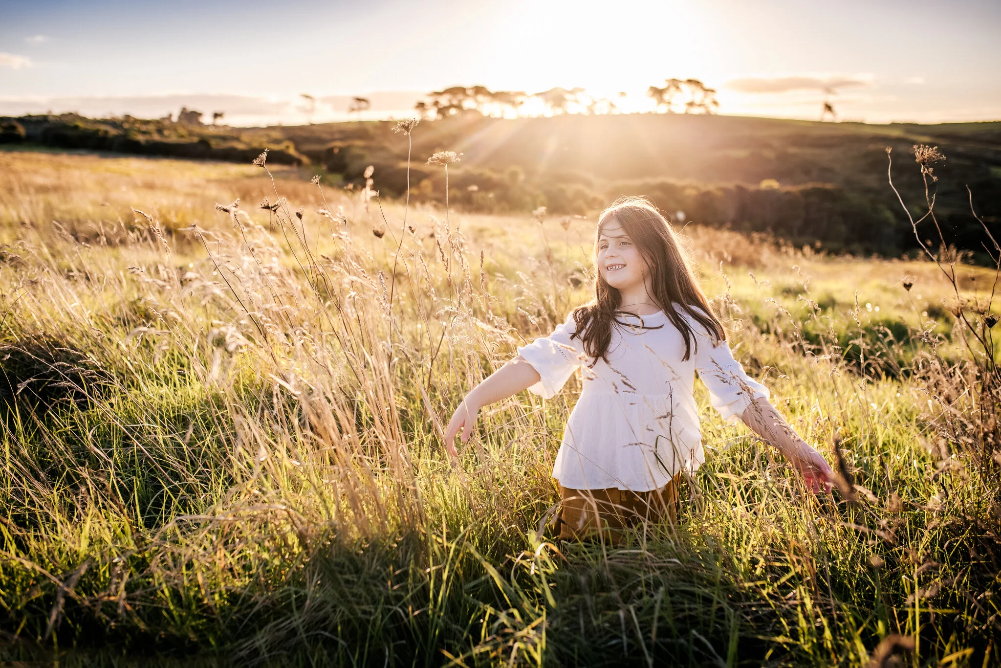 A young girl with long brown hair, wearing a white dress, smiling and playing in a field of tall grass with the sun setting behind her.