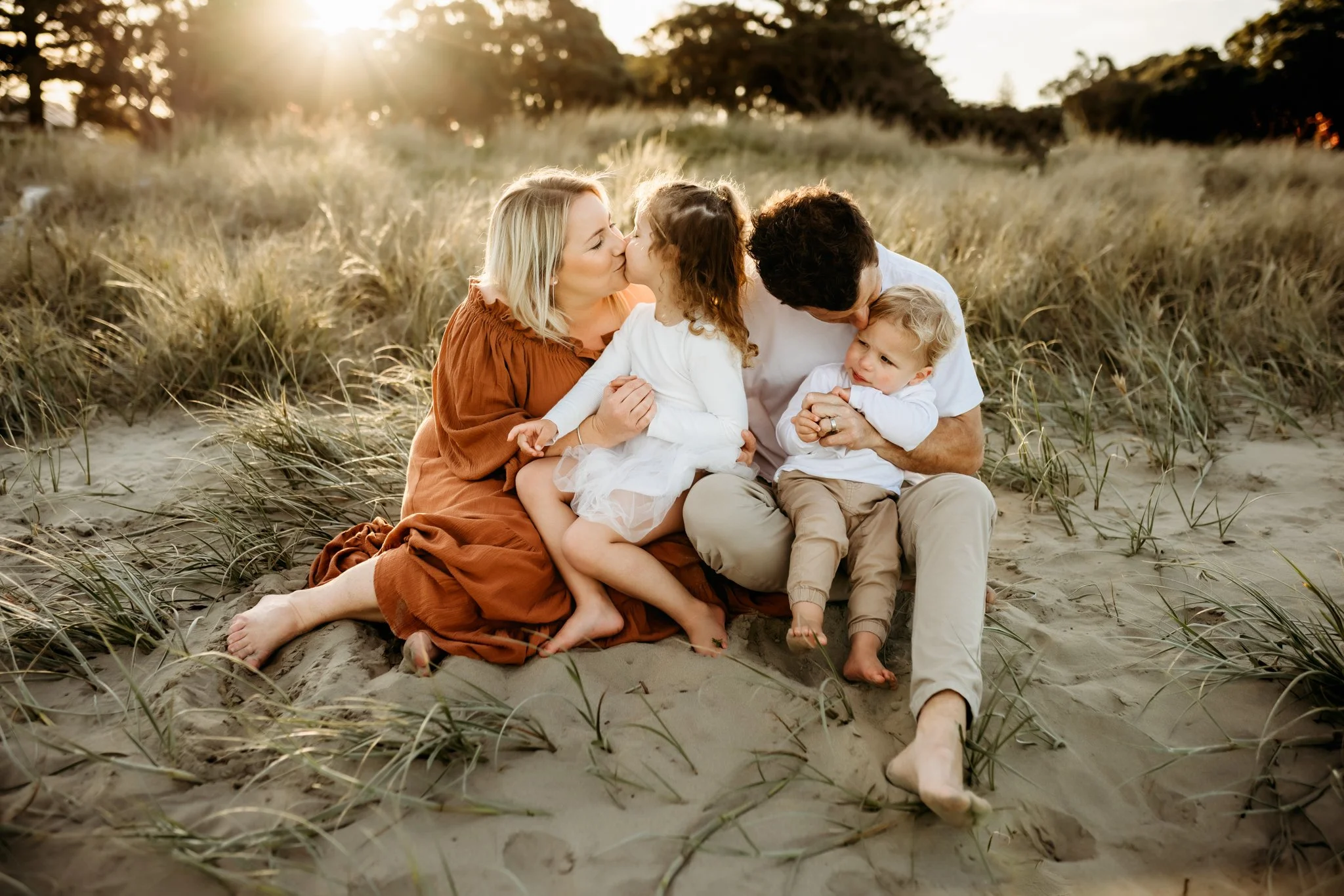 A family of four sitting on the sand at a beach during sunset, sharing a kiss and hugging each other, with tall grasses around them and trees in the background.