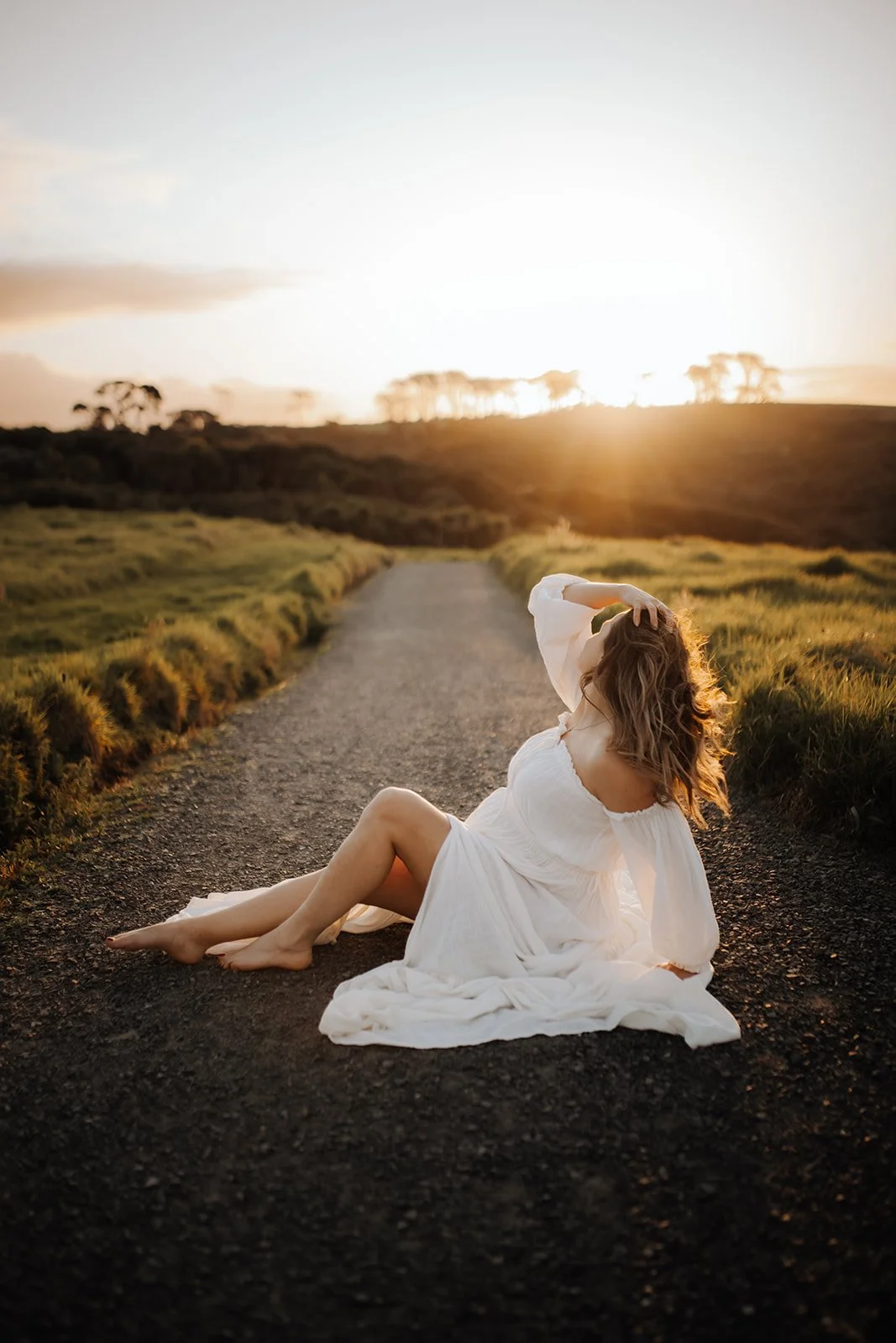 A woman in a white dress sitting on a rural dirt road at sunset, with one hand on her head and her hair blowing in the breeze.