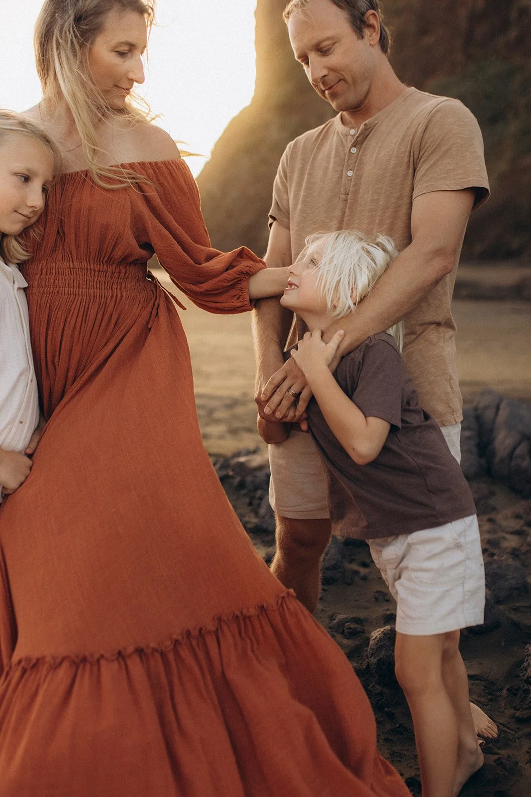 A family of four, including a woman, a man, and two young children, on a beach at sunset. The woman is touching the woman's belly while the children look on with affection. The scene is warm and tender.