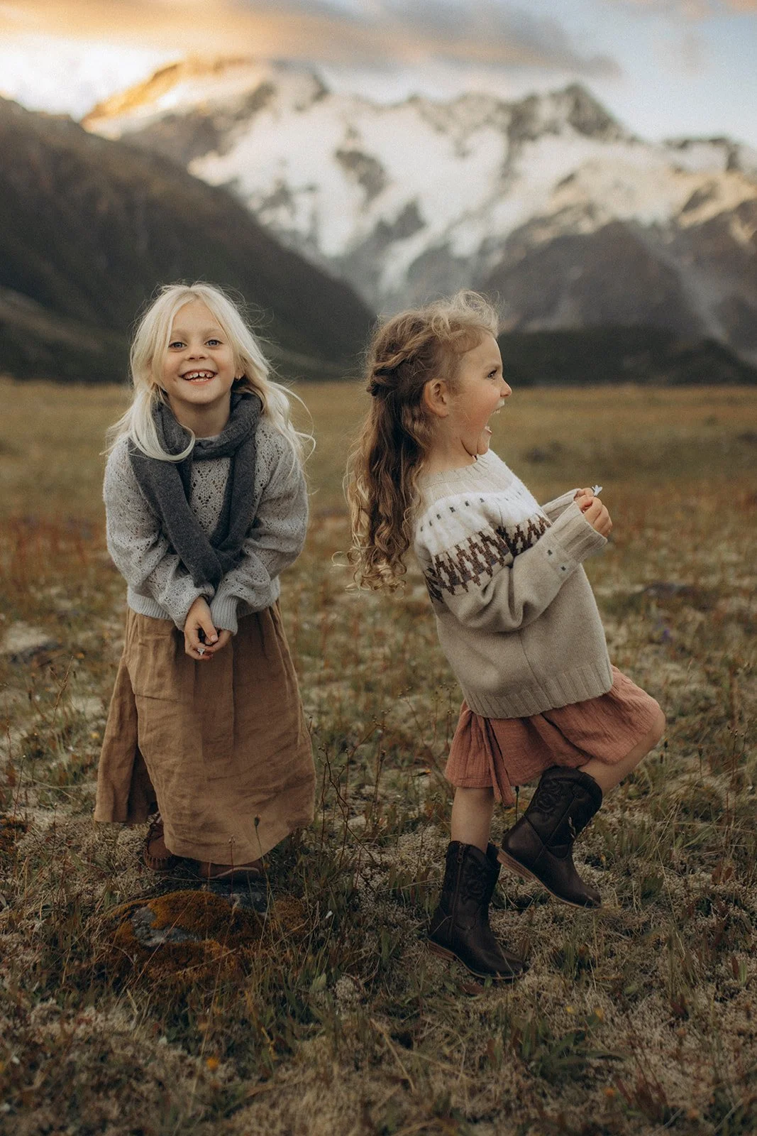 Two young girls laughing and playing outdoors in a grassy field with mountains in the background.