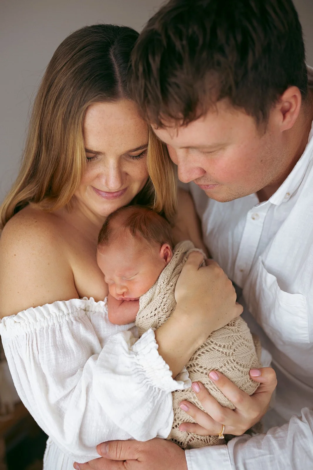 A woman holding a newborn baby, with a man leaning in close, all smiling softly in a tender moment.
