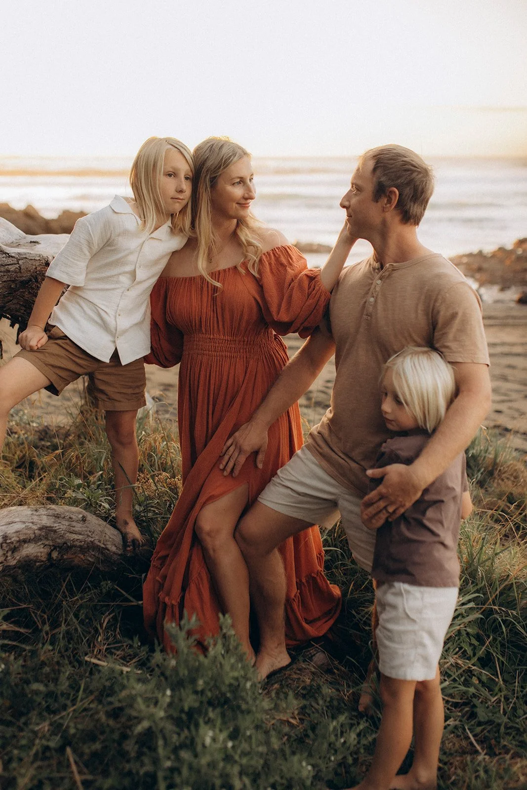 A family of five at the beach during sunset, with a woman in a long orange dress, two young boys, and two young girls.