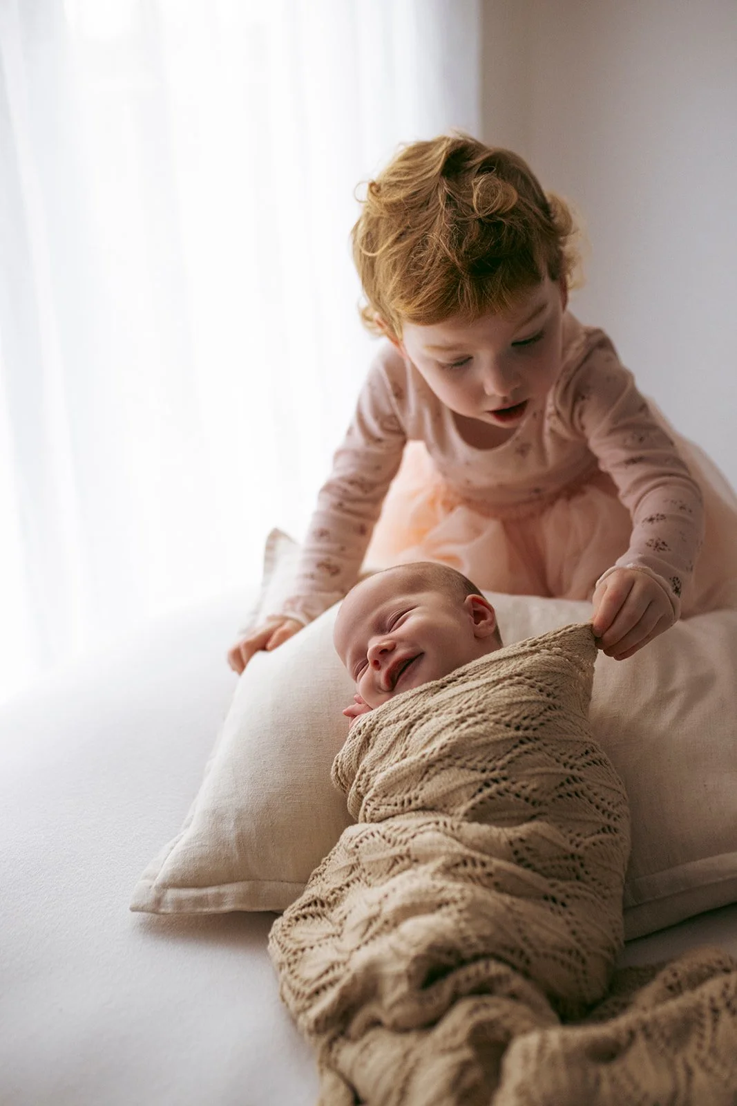A young girl with curly blonde hair playing with a smiling baby wrapped in a beige blanket on a bed near a window with sheer curtains.