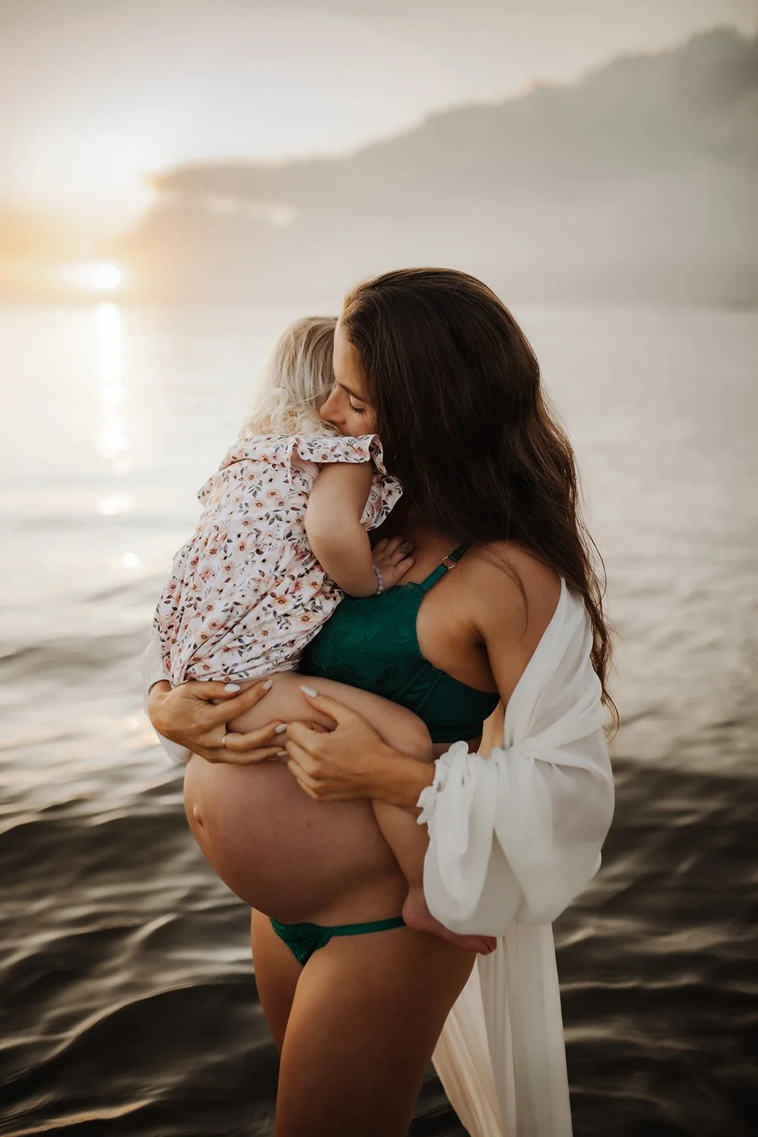 A woman in a green bra and white cover-up holding a young girl, likely a mother and daughter, on a beach during sunset.