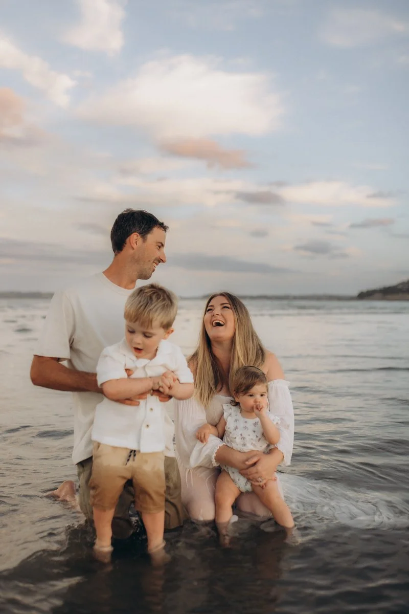 A family of four enjoying time together at the beach during sunset, with the parents smiling and two young children, one standing and the other sitting in the shallow water.
