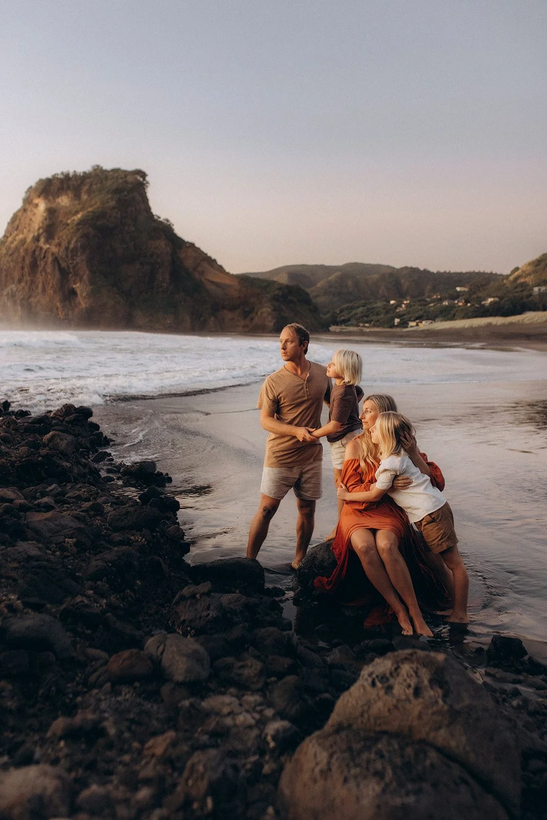 Family of five on rocks by the ocean with a large hill in the background, during sunset or sunrise.