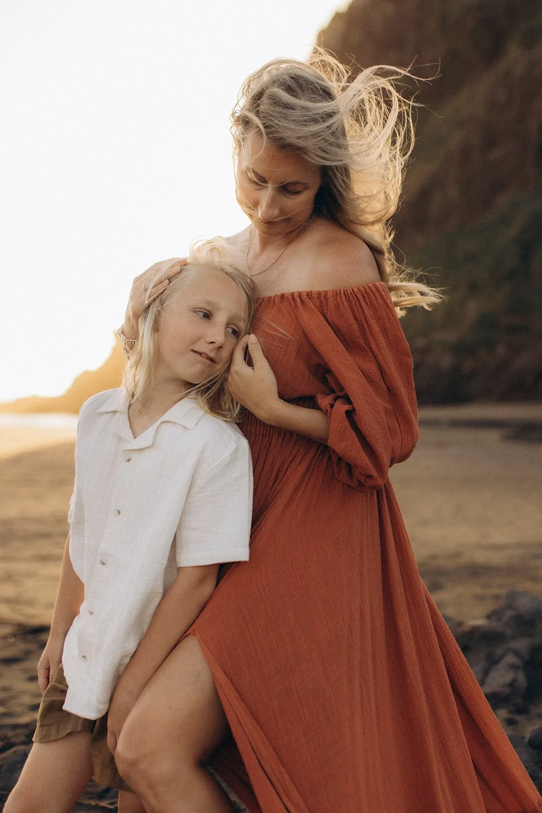 A woman with long blonde hair in an orange off-shoulder dress hugging a young girl with blonde hair in a white shirt on a beach at sunset.