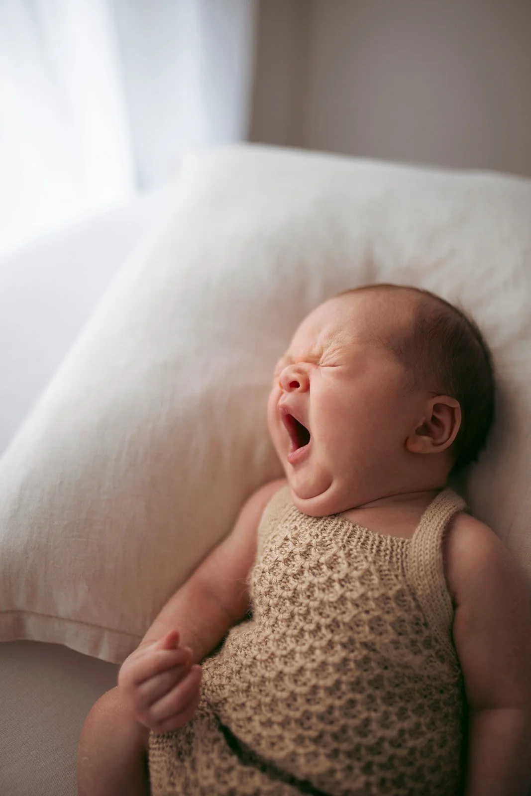 A baby lying on a cushion yawning with eyes closed and mouth wide open.