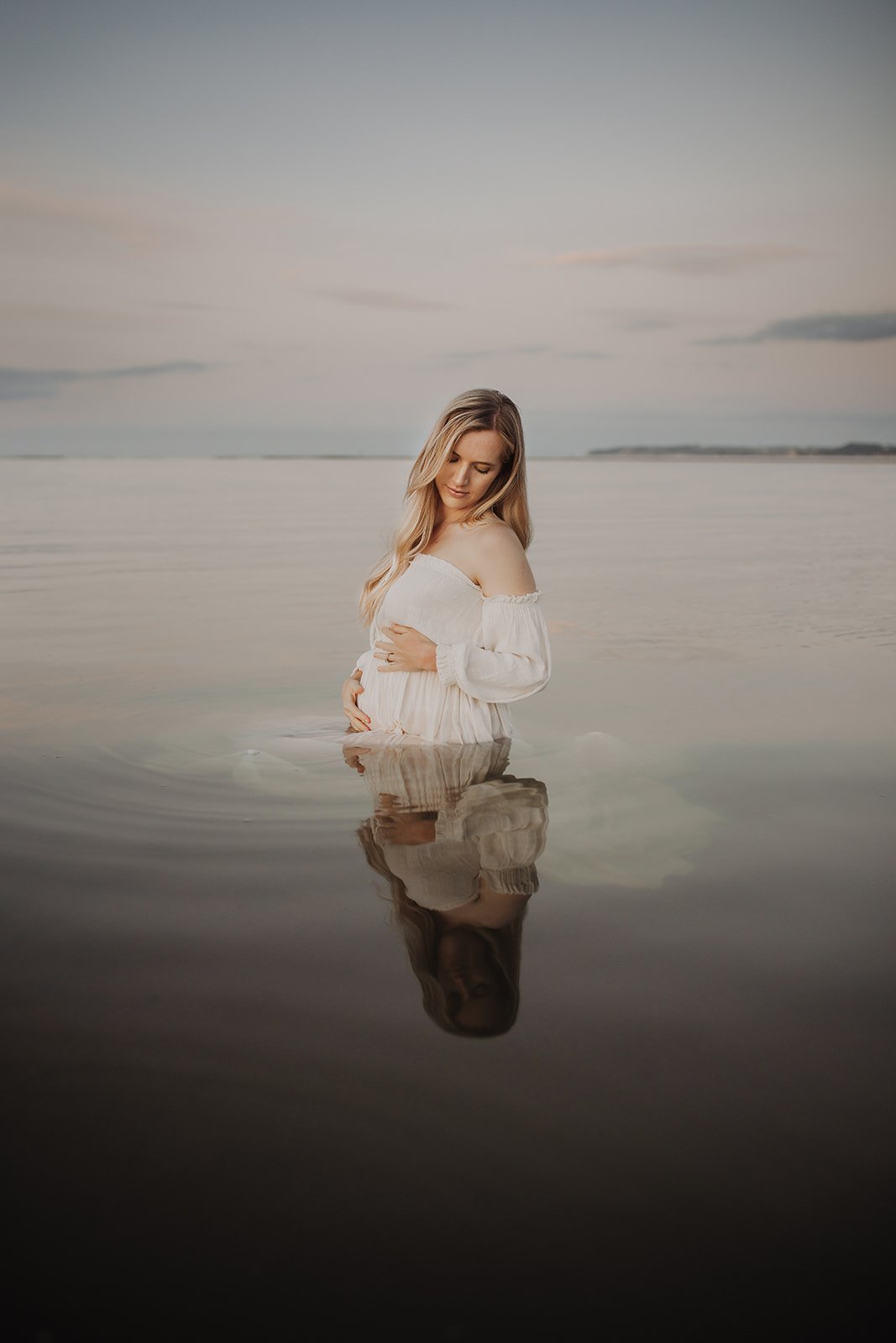A pregnant woman standing in shallow water at the beach during sunset with her hand on her belly and looking down, wearing a white off-shoulder dress, with her reflection visible in the water.