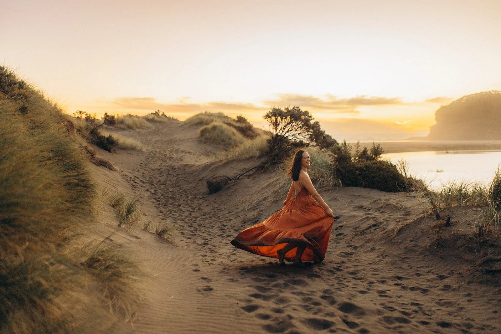 A woman in an orange dress walking on sandy dunes at sunset.