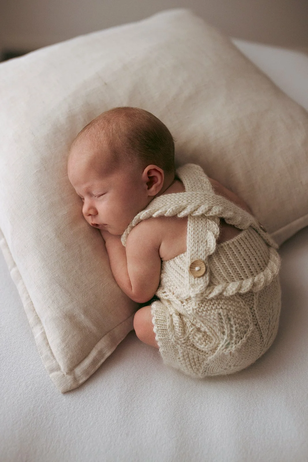 A sleeping baby curled up on a beige pillow, wearing a knitted cream-colored outfit with a button detail.