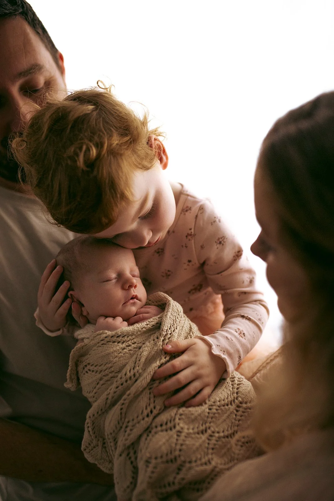 A family scene with a woman holding a newborn baby wrapped in a knitted blanket, a young girl kissing the baby's forehead, and a man smiling nearby, all sharing a tender moment.