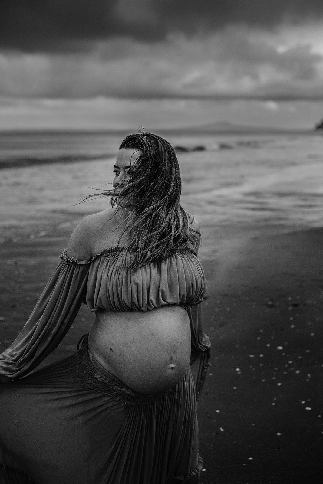 Black and white photo of a woman sitting on the beach, looking over her shoulder, with the ocean and cloudy sky in the background.