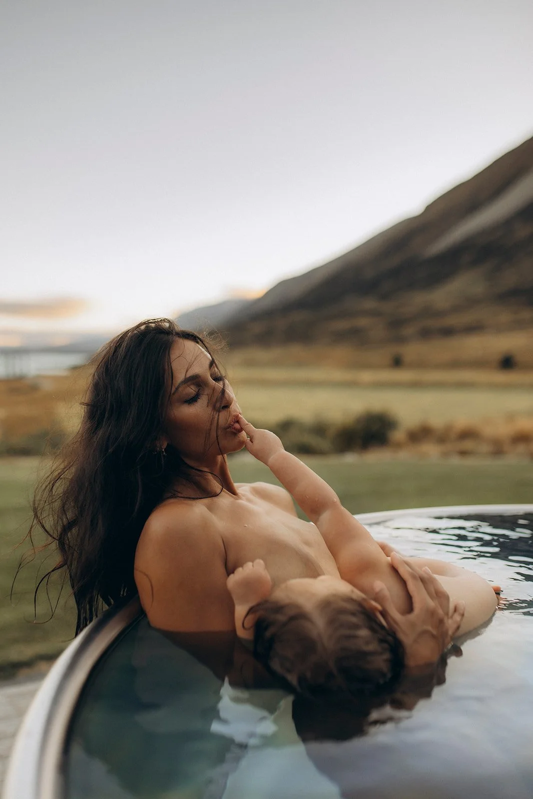 A woman with long dark hair relaxing in a hot tub outdoors with a young child, beautiful landscape with hills and a cloudy sky in the background.