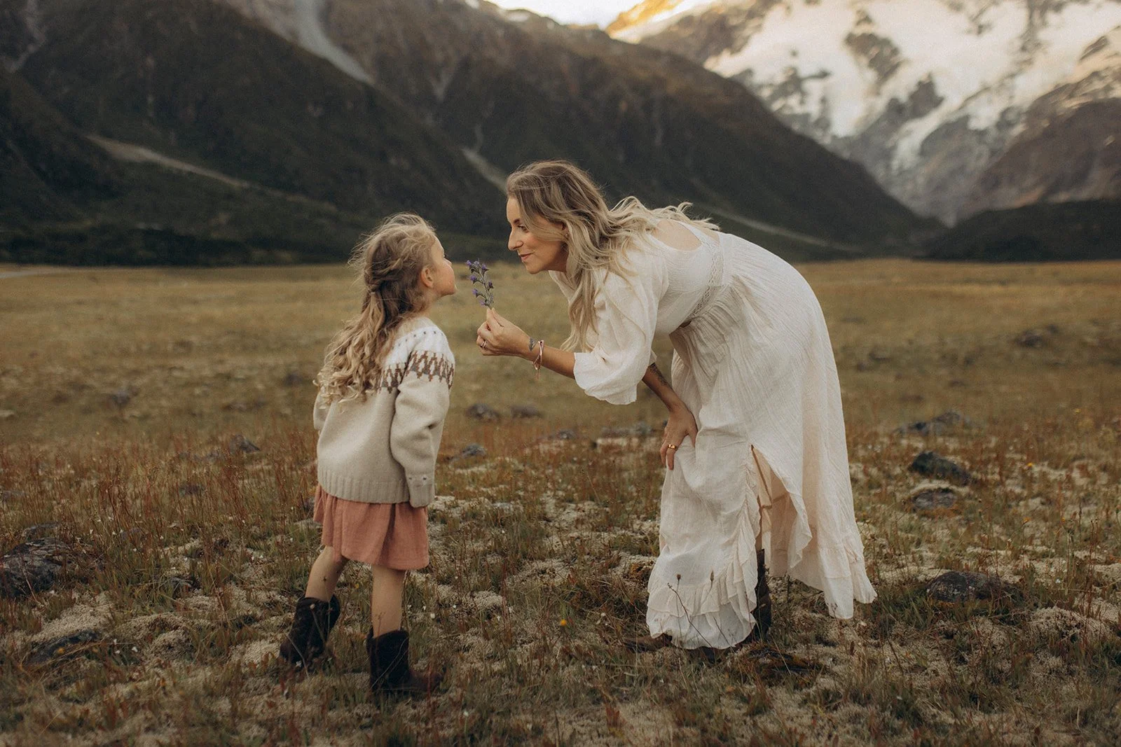 A woman with long blonde hair wearing a white flowing dress is bending over, holding a purple flower close to a young girl with curly blonde hair, who is wearing a beige sweater, pink skirt, and black boots, in a mountain field with snow-covered peak