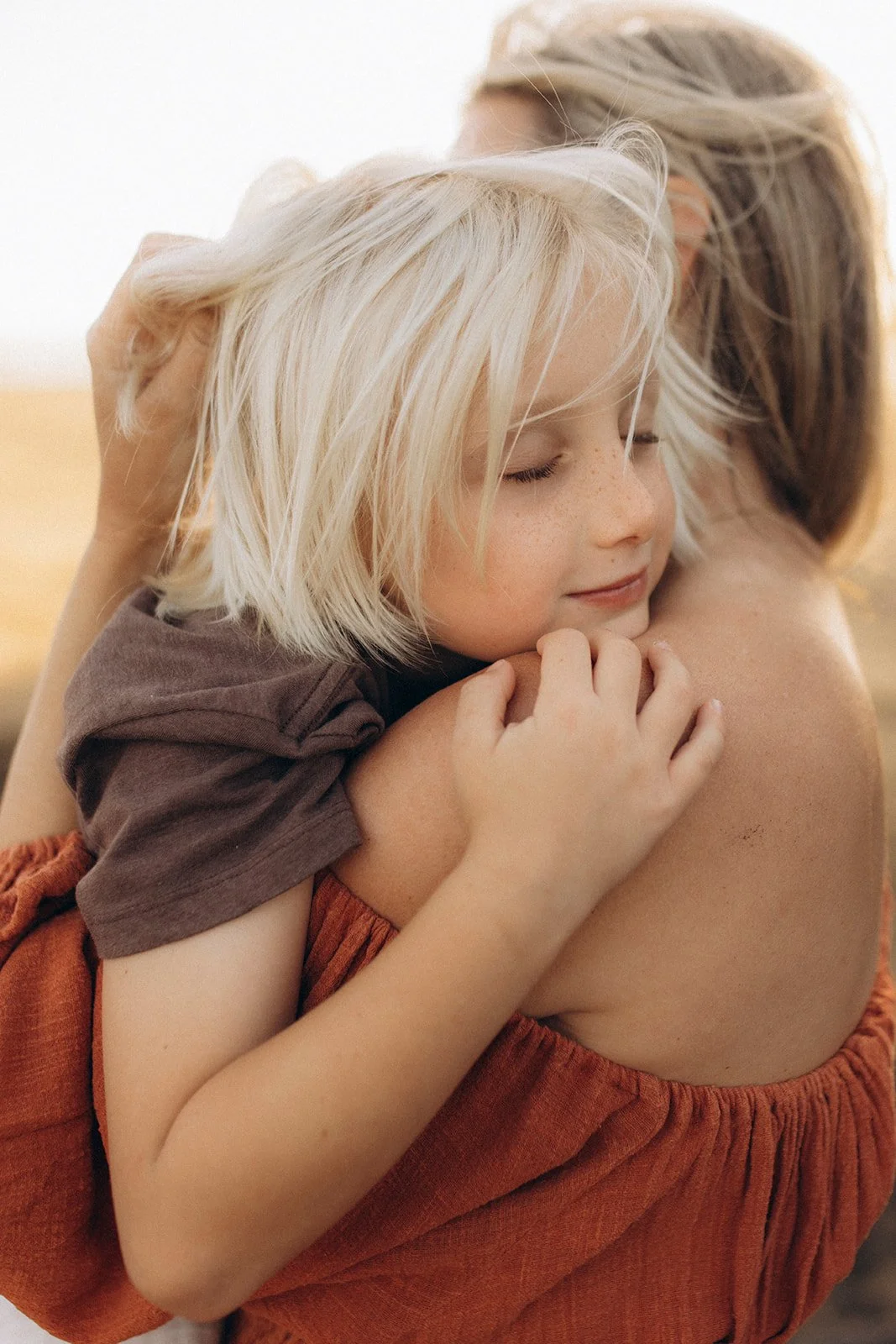 A young girl with blonde hair hugging an adult person, possibly her mother, with both of them having their eyes closed and appearing content.