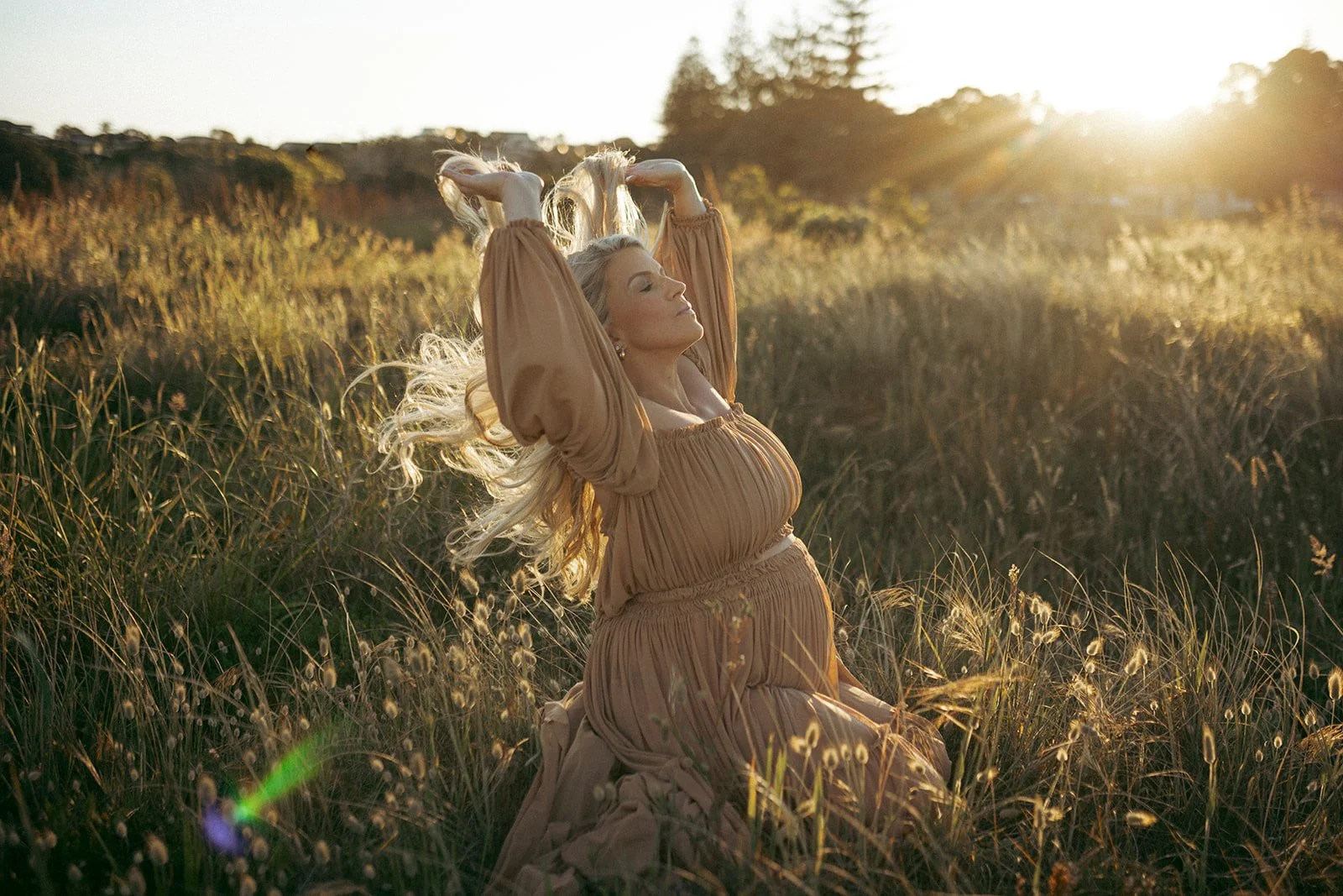 A woman with long blond hair in a flowing beige dress kneels in a grassy field at sunset, with her eyes closed and arms raised above her head, embracing the sunlight.