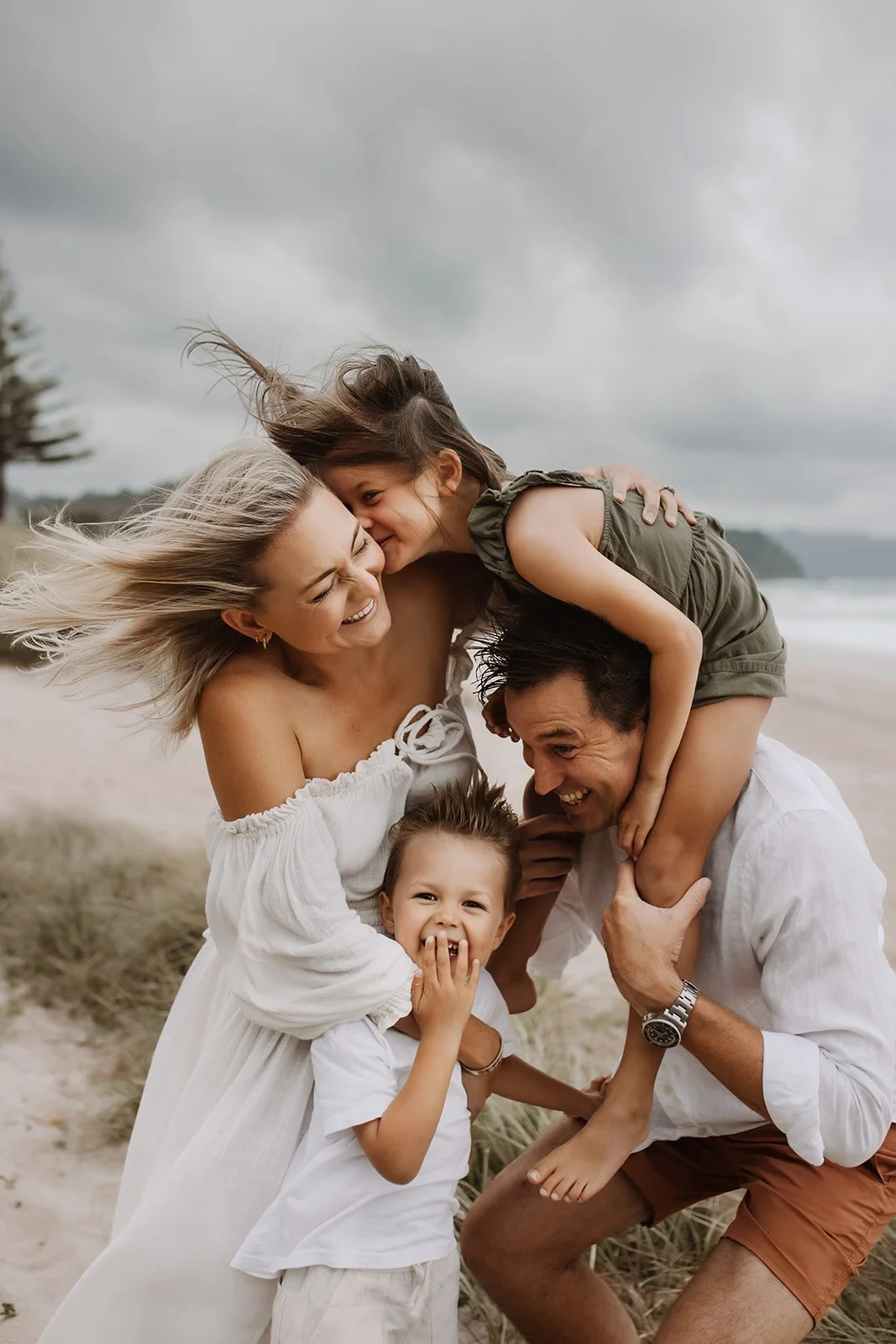 Family of four enjoying time on a beach, playful and smiling as they interact with each other under cloudy skies.