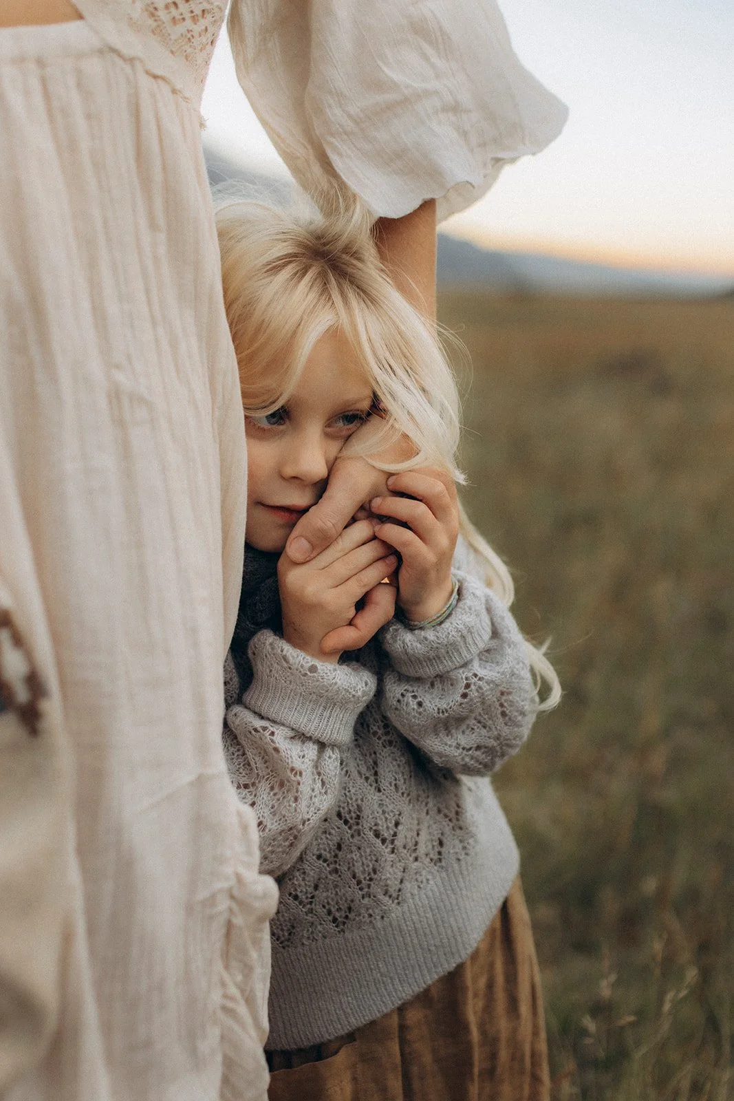 A young girl with blonde hair hugging an adult, likely her mother, outside during sunset or late afternoon, with a blurred natural background.