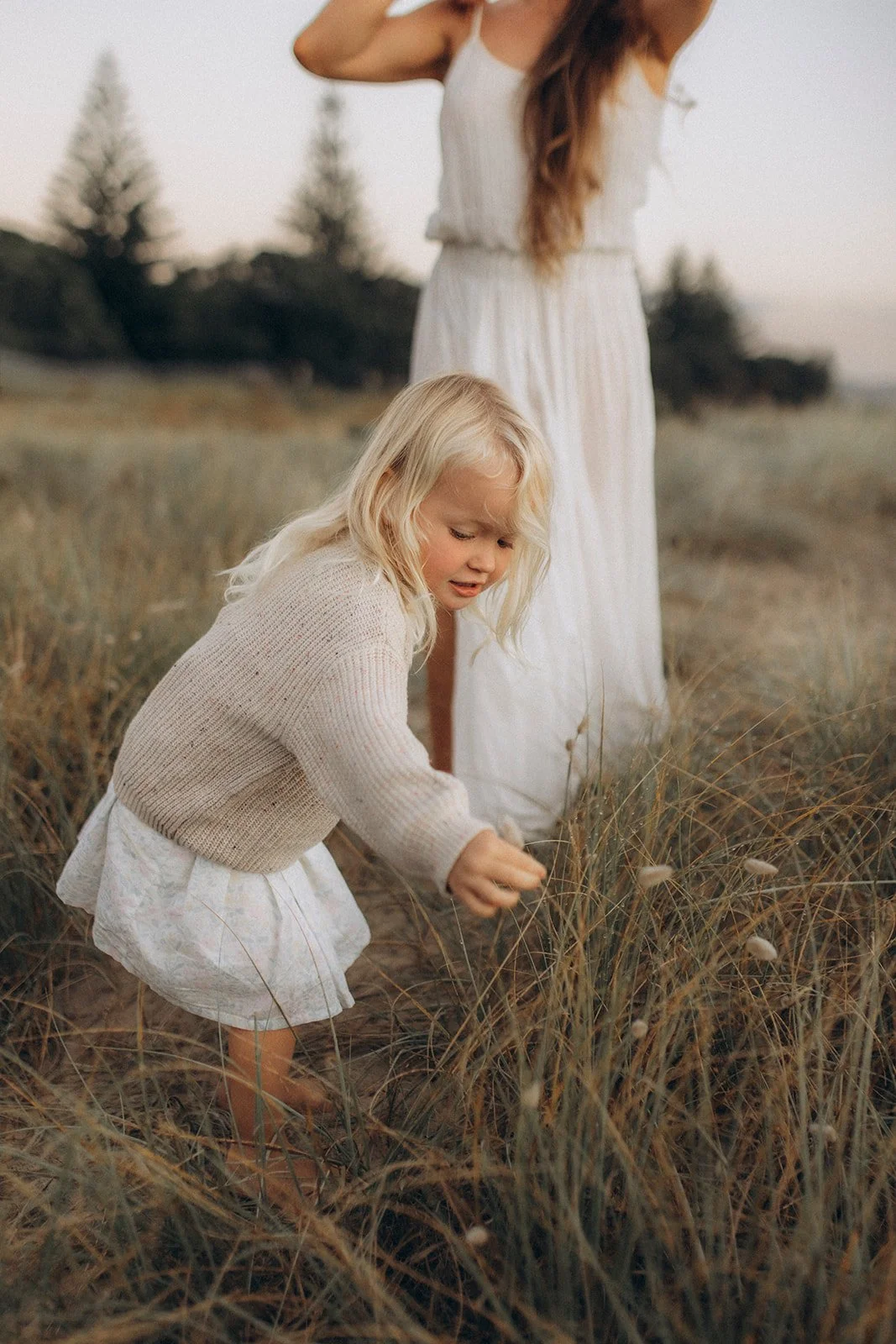 A young girl with blonde hair wearing a knitted sweater and a white dress, crouching in a field of tall grass, with a woman standing behind her, both in light-colored dresses, during dusk or early evening.