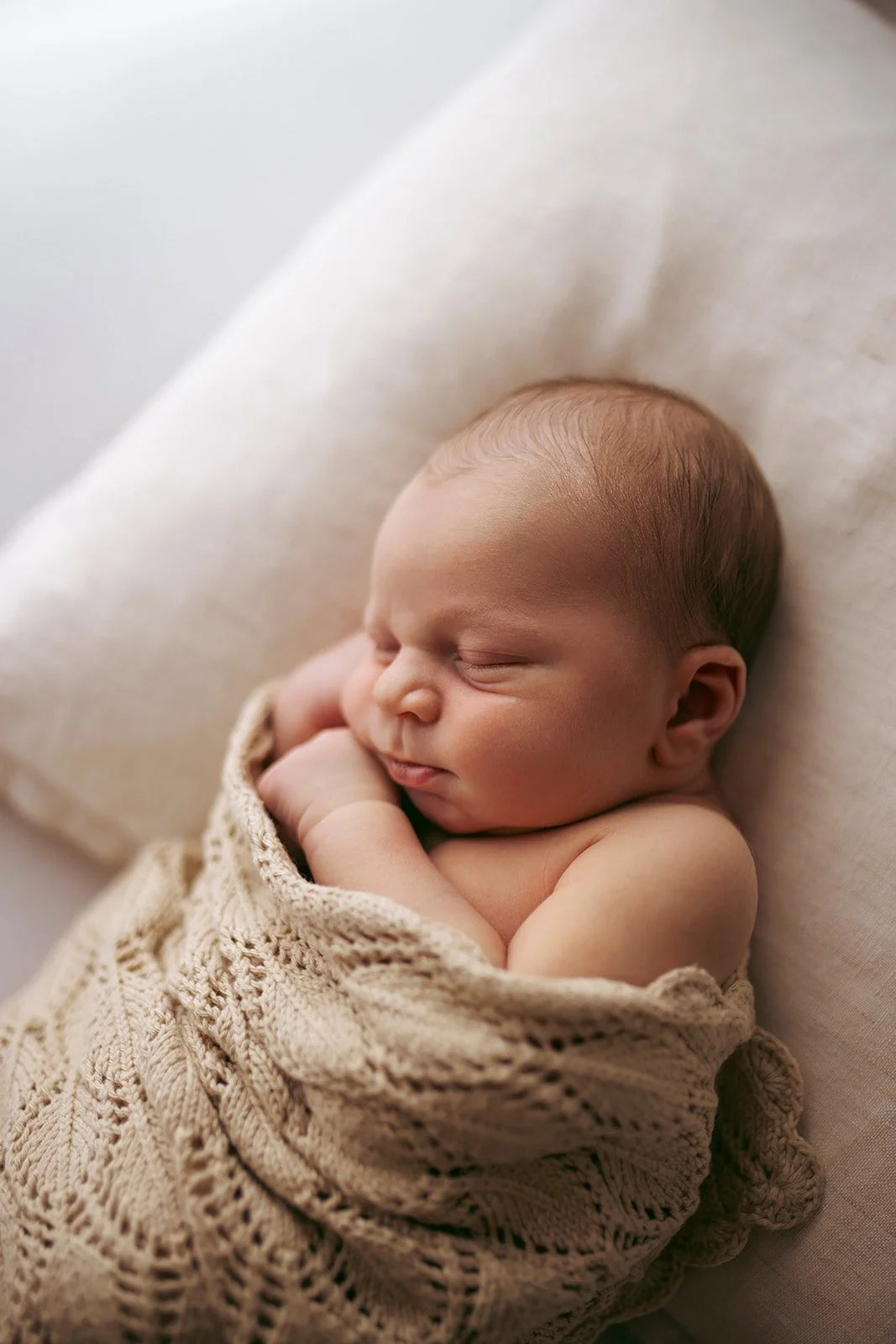A sleeping newborn baby wrapped in a beige knit blanket, lying on a light-colored cushioned surface.